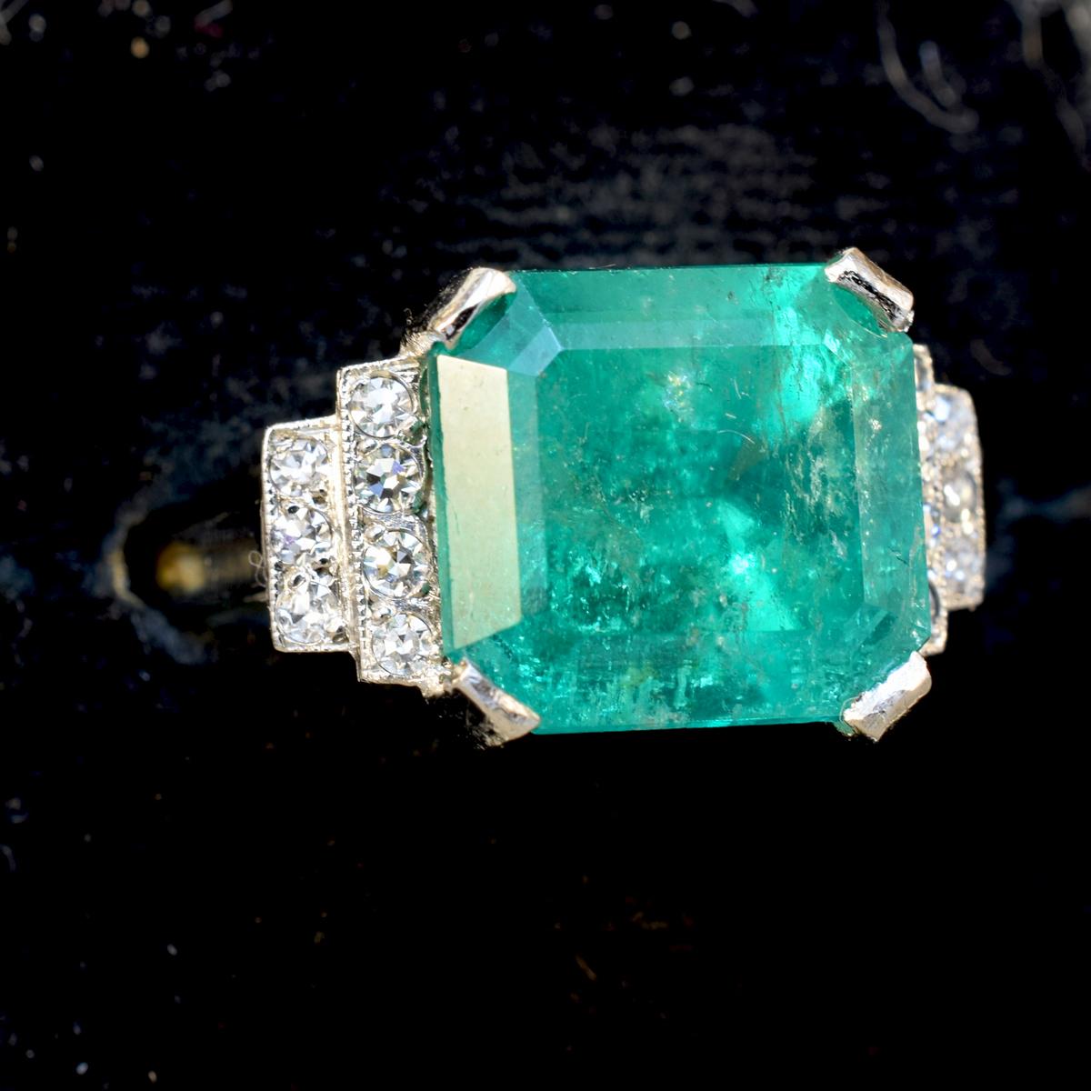 An Emerald single stone ring with a Diamond set stepped down shoulders. The Emerald Cut Emerald weighs 5.22ct in a four claw corner cut setting to an openwork mount, rectangular stepped down graduating shoulders, grain set with a total of fourteen