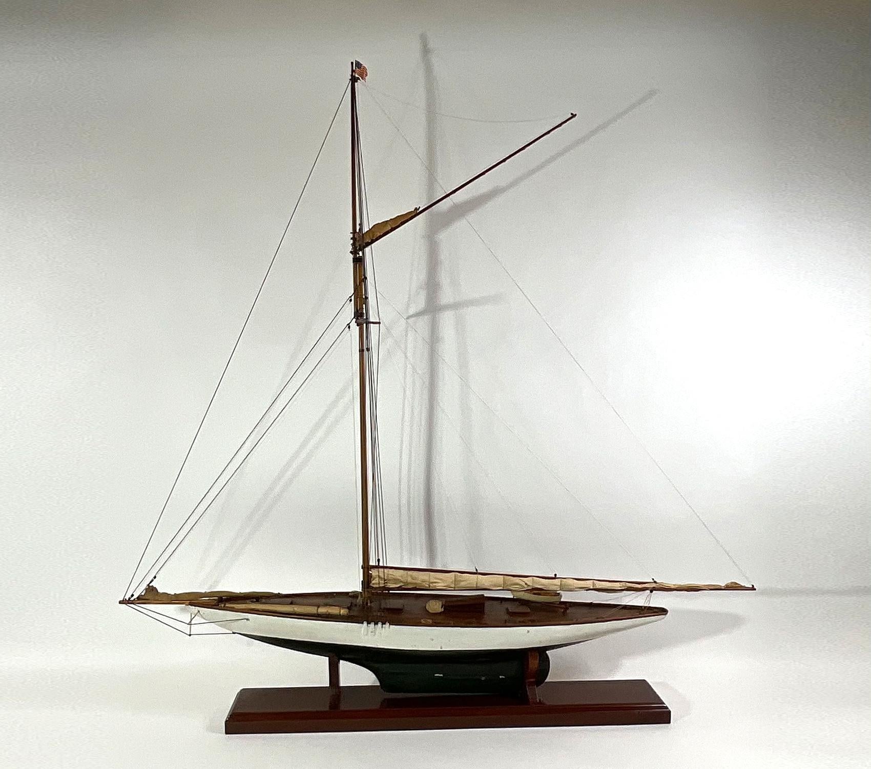 Antique yacht model with varnished deck, cabins, hatches, launch on davits. Hull is fitted with a lead keel. Hull is painted green below the waterline and white above. Furled sail is lashed to the boom. Two more sails are lashed to the bow and