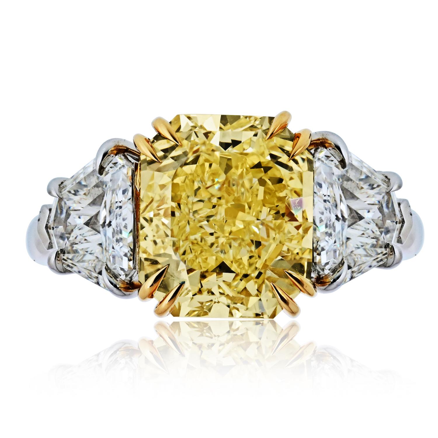 Questo squisito anello di fidanzamento con tre pietre è un capolavoro di eleganza senza tempo e di raffinata maestria. Al centro brilla un diamante Fancy Yellow da 5,47 carati dal taglio radiante, certificato GIA e classificato VVS2 per la sua