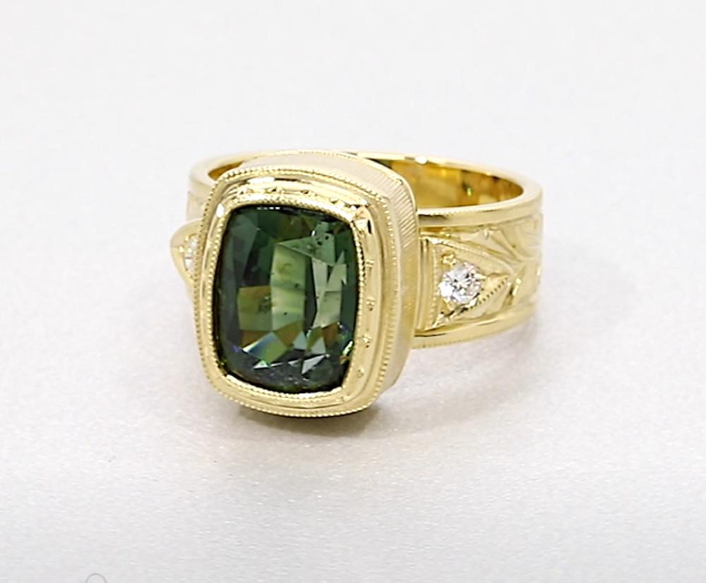 This gorgeous handmade 18k yellow gold ring features a 5.49 carat cushion-cut green tourmaline with color as lush as a tropical rainforest. Intricate engraving around the entire bezel is visible from every angle, and two round brilliant cut diamonds