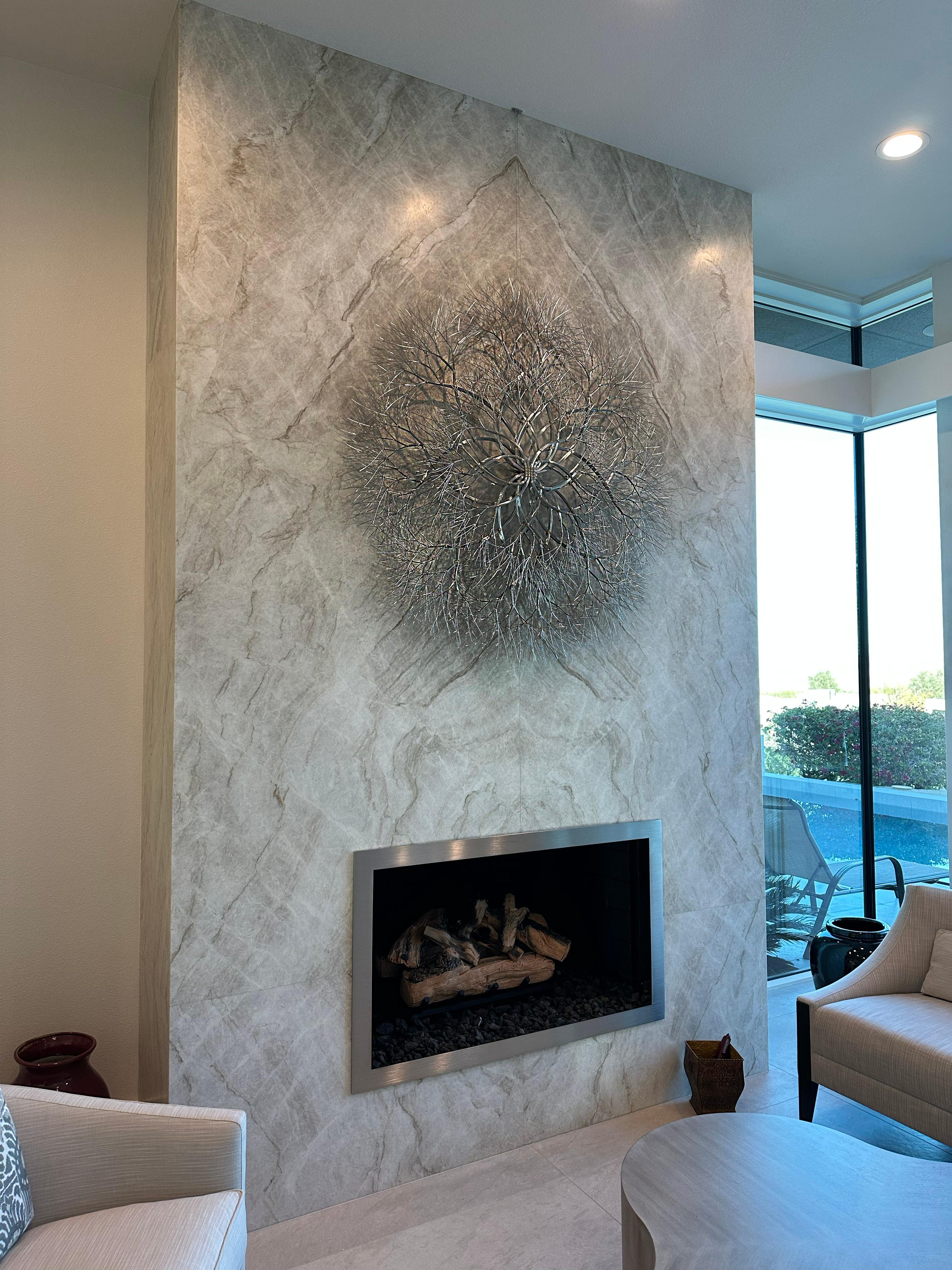 Stainless steel. 

Kue King created this elongated wall mounted sculpture using stainless steel wire. The polished stainless steel gives the sculpture a shine, reflectivity, and mirrored finish that adds sparkle to the room and picks up on colors