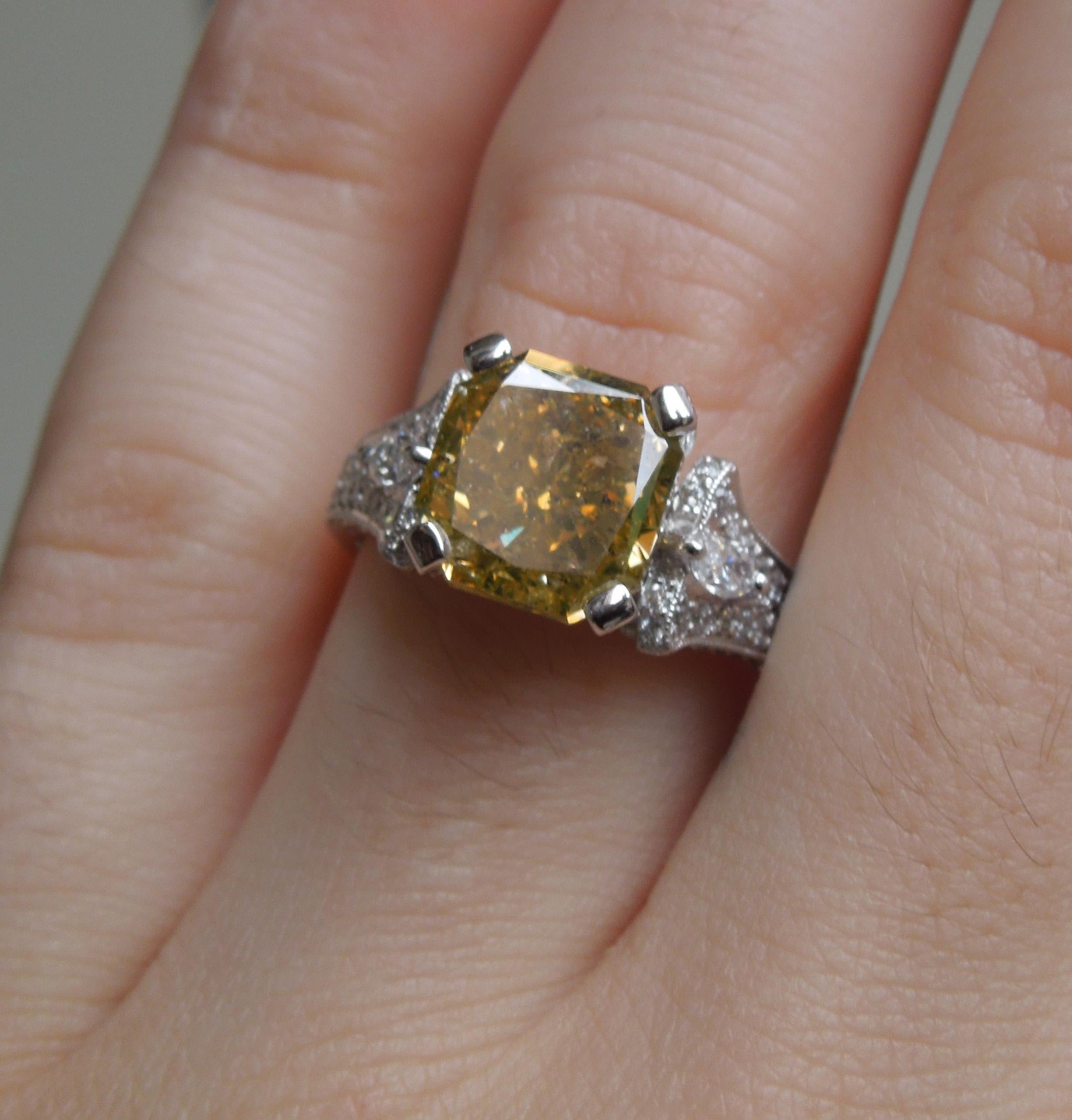 Constructed of 18 Karat White Gold, featuring a focal Asscher cut Natural Golden Canary [Zimi] Diamond weighing approximately 5.50 carats ranking an I1 Clarity, measuring 9.5mm x 9.1mm, securely Prong-set. 

Accented with 2 Side Marquise cut