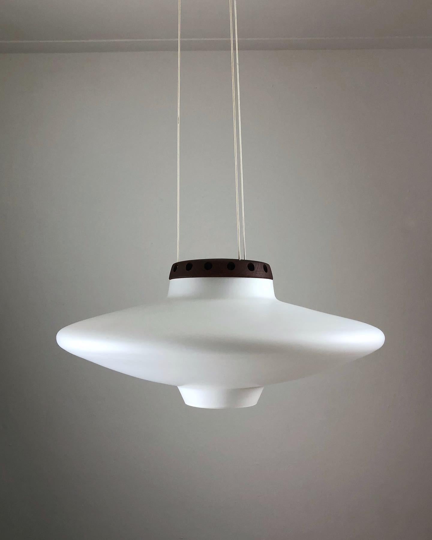 The shape of this large lamp is very similar to a ufo. Designed by the Swedish brothers Uno & Östen Kristiansson and manufactured by the furniture company Luxus Vittsjö in the 1950s.
The lamp is made of white matte opal glass and teak wood. These 2