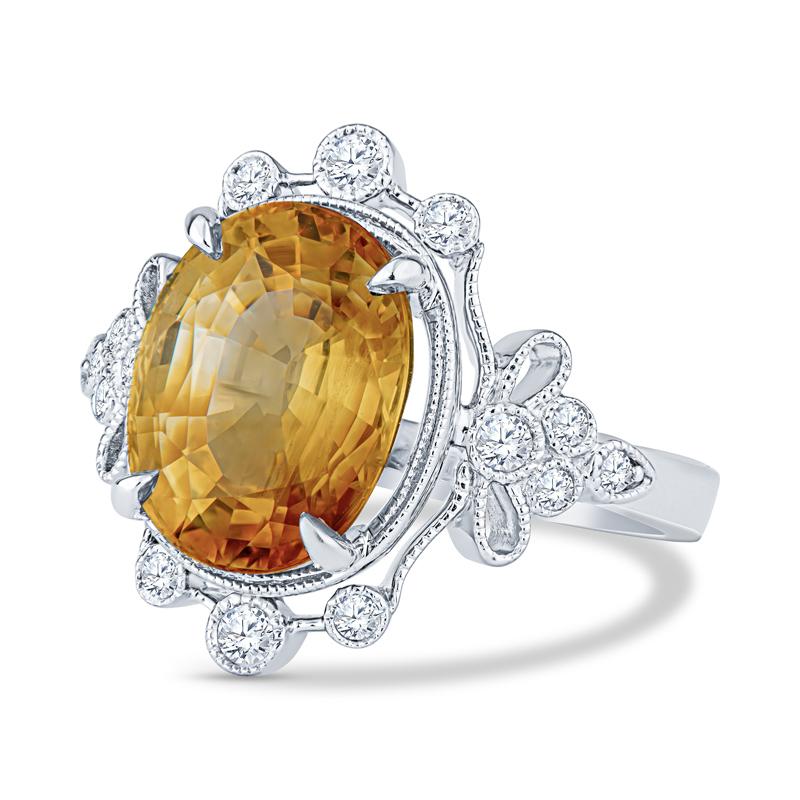 This vintage inspired ring features a 5.70 carat orange-yellow oval cut sapphire accented by 0.20 carat total weight in round diamonds with milgrain details set in 14 karat white gold. This ring is currently a size 6 but can be resized upon request.