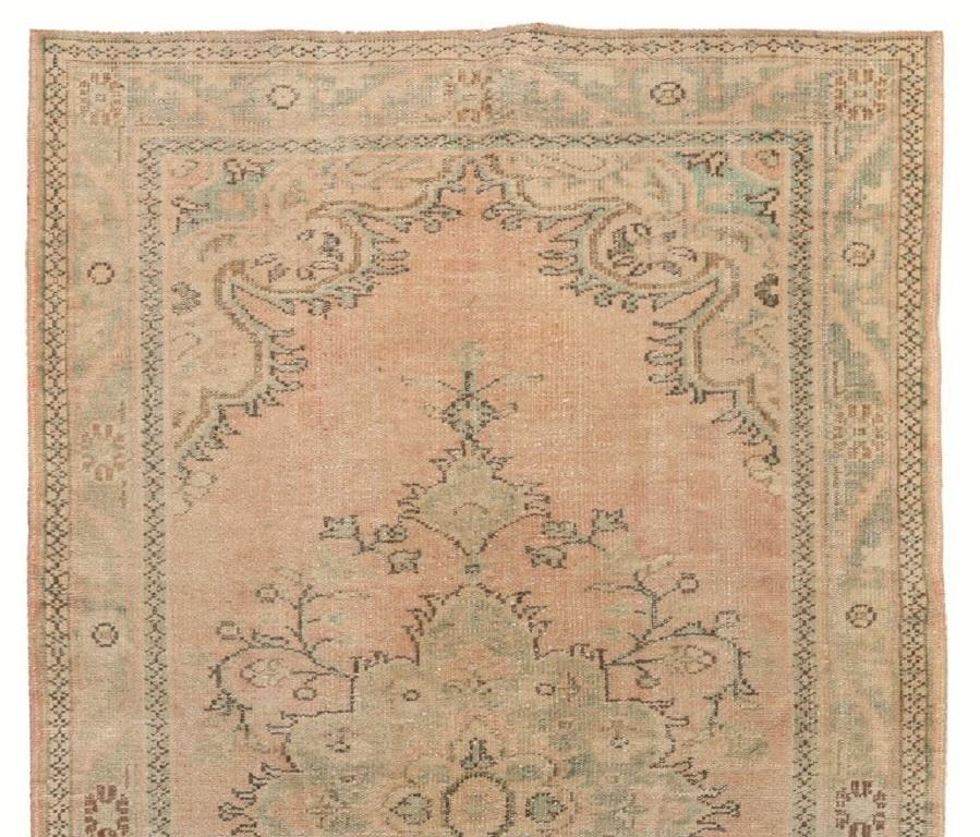 This is a vintage hand-knotted Turkish Oushak area rug featuring a central medallion in soft muted sage green decorated all around with floral vines against a faded red background as well as a border filled with floral heads and serrated leaves. The