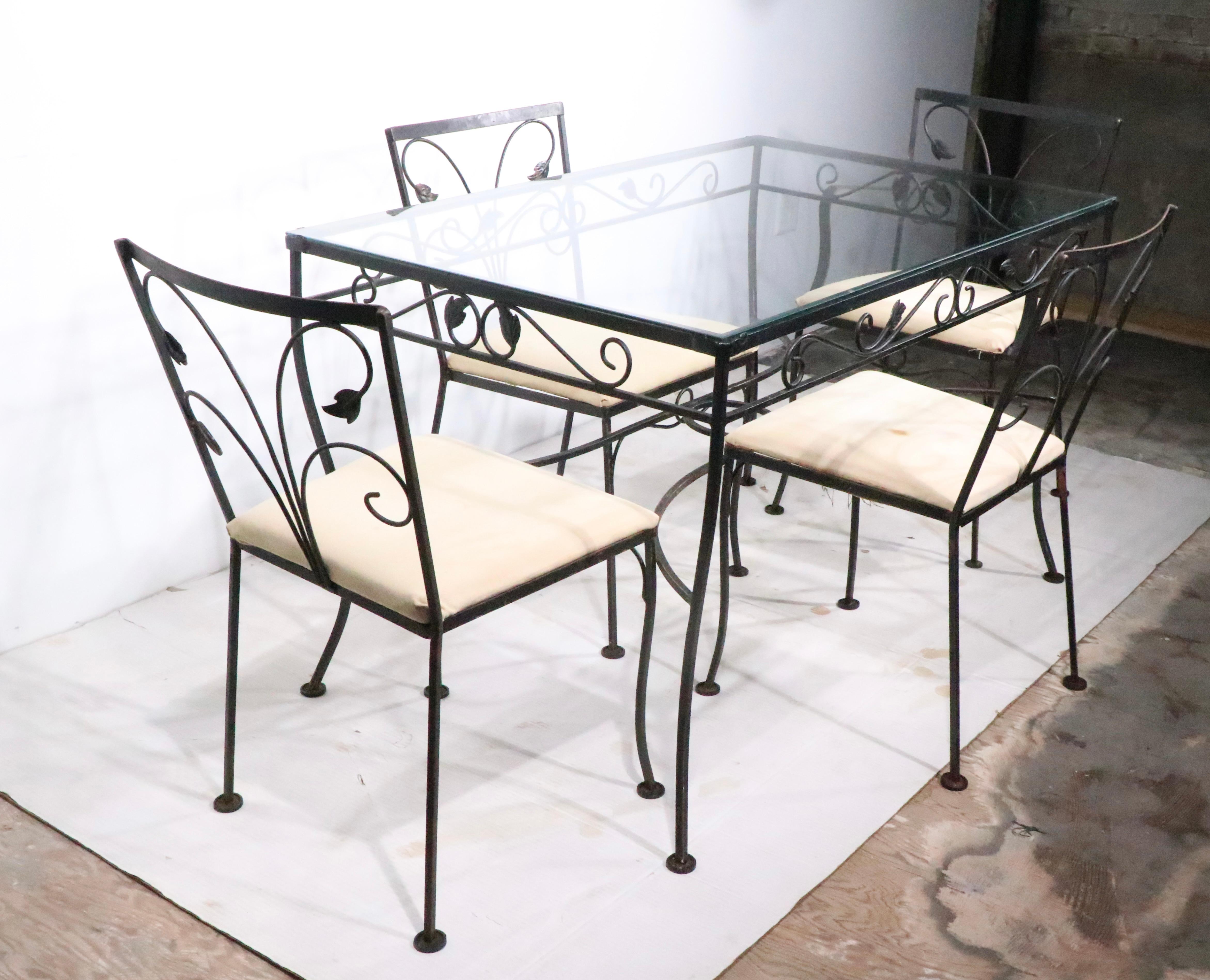 Voguish wrought iron  garden,  patio set including a glass top table, and  four chairs, one arm, three sides. Well crafted, and in very good, clean condition, seat cushions should be recovered.  Structurally sound and sturdy, finish shows some