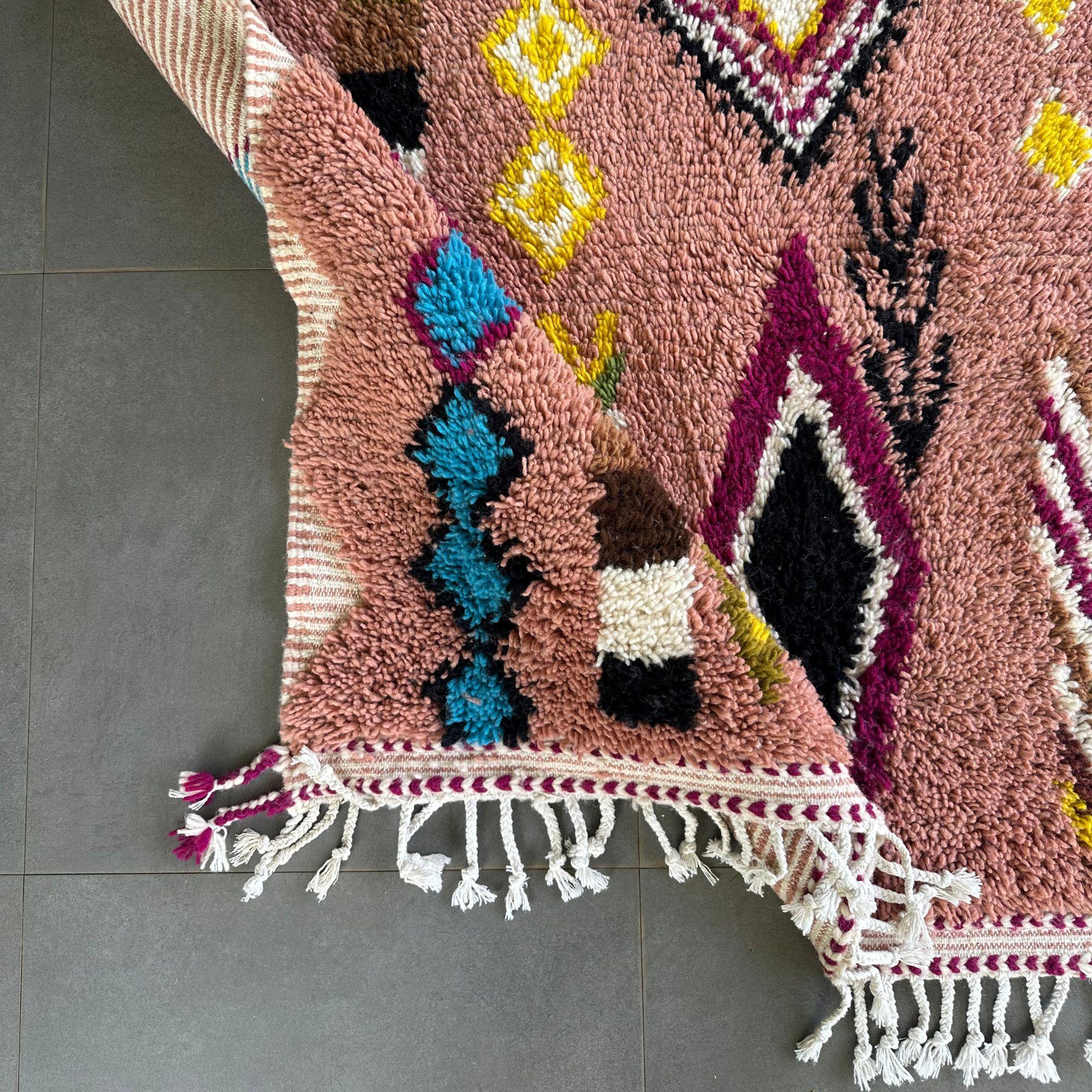 The Artisan Treasures is a vibrant pink Moroccan rug that beautifully merges traditional Berber craftsmanship with modern flair. This handwoven rug features colorful diamond-shaped tribal designs across its soft pink base, along with textured sides