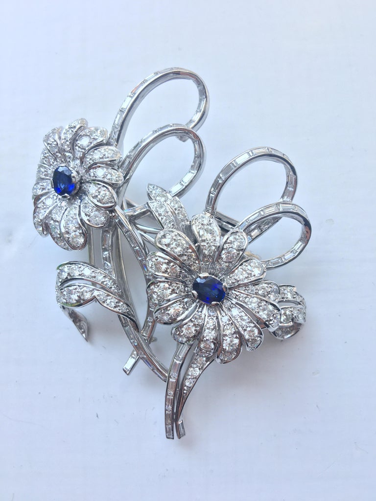 Double Flower shape brooch featuring an oval blue sapphire on each in the centre flanked by various size petals set with superb white round brilliant cut diamonds in various sizes set in 18 karat white gold. The Lustrous white gold diamonds and