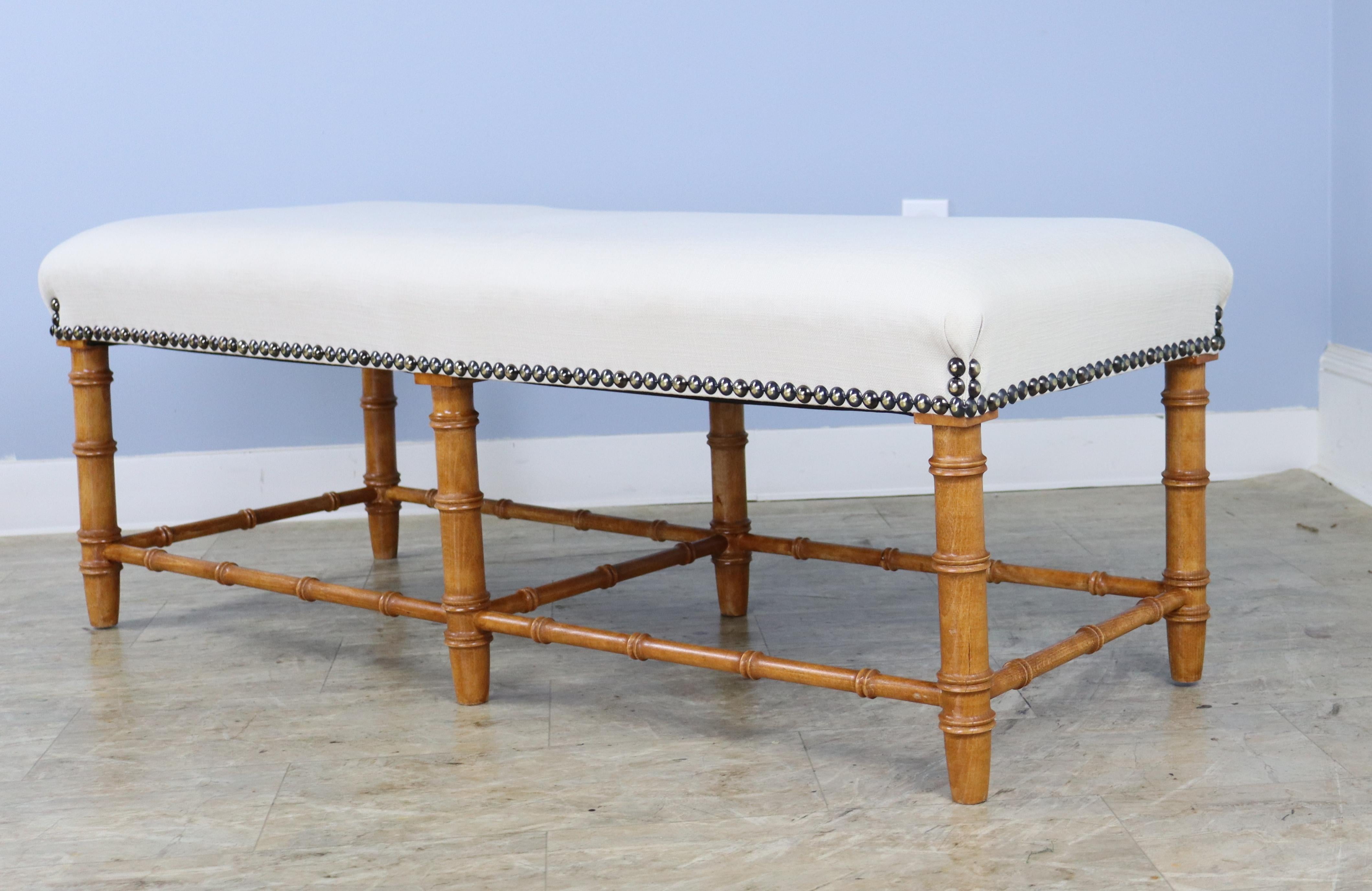 A long comfortable bench or stool in elegant faux bamboo, possibly carved of English Golden Oak. A classic look, newly uphostered in cream linen.  If you are looking for a pair, we also have 0324-M105B available.
