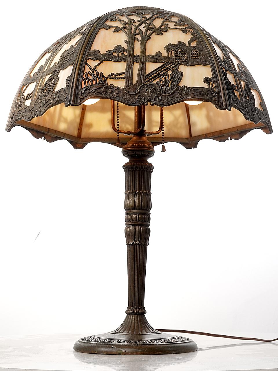 This is a nicely designed Arts and Crafts table lamp. It has a curved shade with 6 elaborate landscape themed cast filigree panels. Each slump glass (curved) panel features a cream -  amber variegated glass and is lit by two E26 bulbs. All the cast