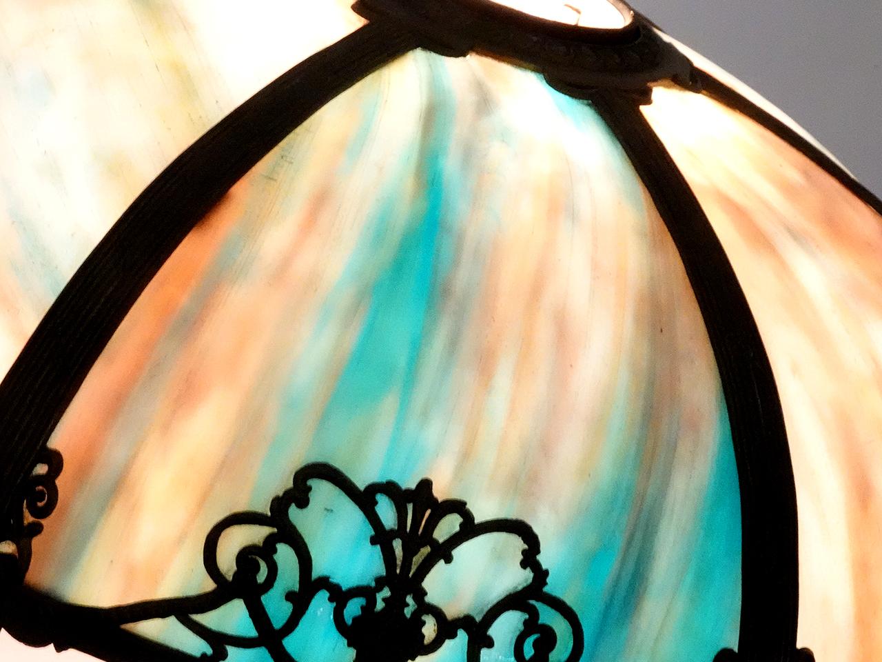 This is a nicely designed Victorian table lamp. It has a curved shade with 6 unique decorative filigree overlays. Each slump glass (curved) panel features a unique blue, cream and amber variegated glass. It is lit by one E26 bulb. All the cast metal