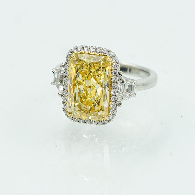 Dieser Ring ist mit einem 6,03-Karat-Diamanten im Radiant-Schliff in der Farbe Gelb besetzt. Die beiden seitlichen Steine bestehen aus insgesamt 0,78 Karat weißen Diamanten und der Halo, der den Mittelstein umgibt, hat insgesamt 0,23 Karat. Der