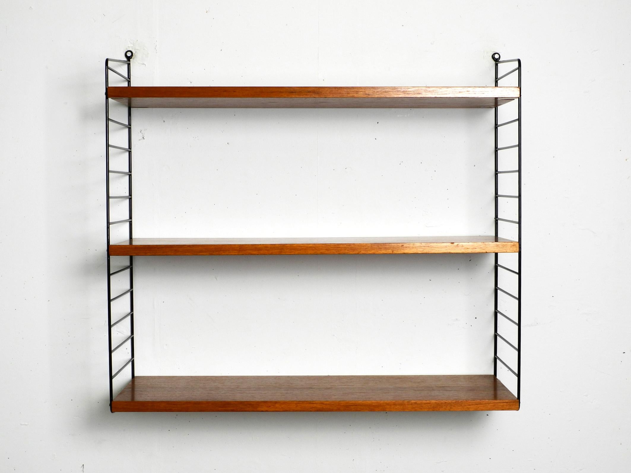 Original 1960s Nisse Strinning wall hanging shelf with three solid wood shelves with teak veneer.
This shelf has the deeper ladder and 30cm deep boards.
2 Metal ladders with black plastic coating.
All two ladders are in good condition. No rust.