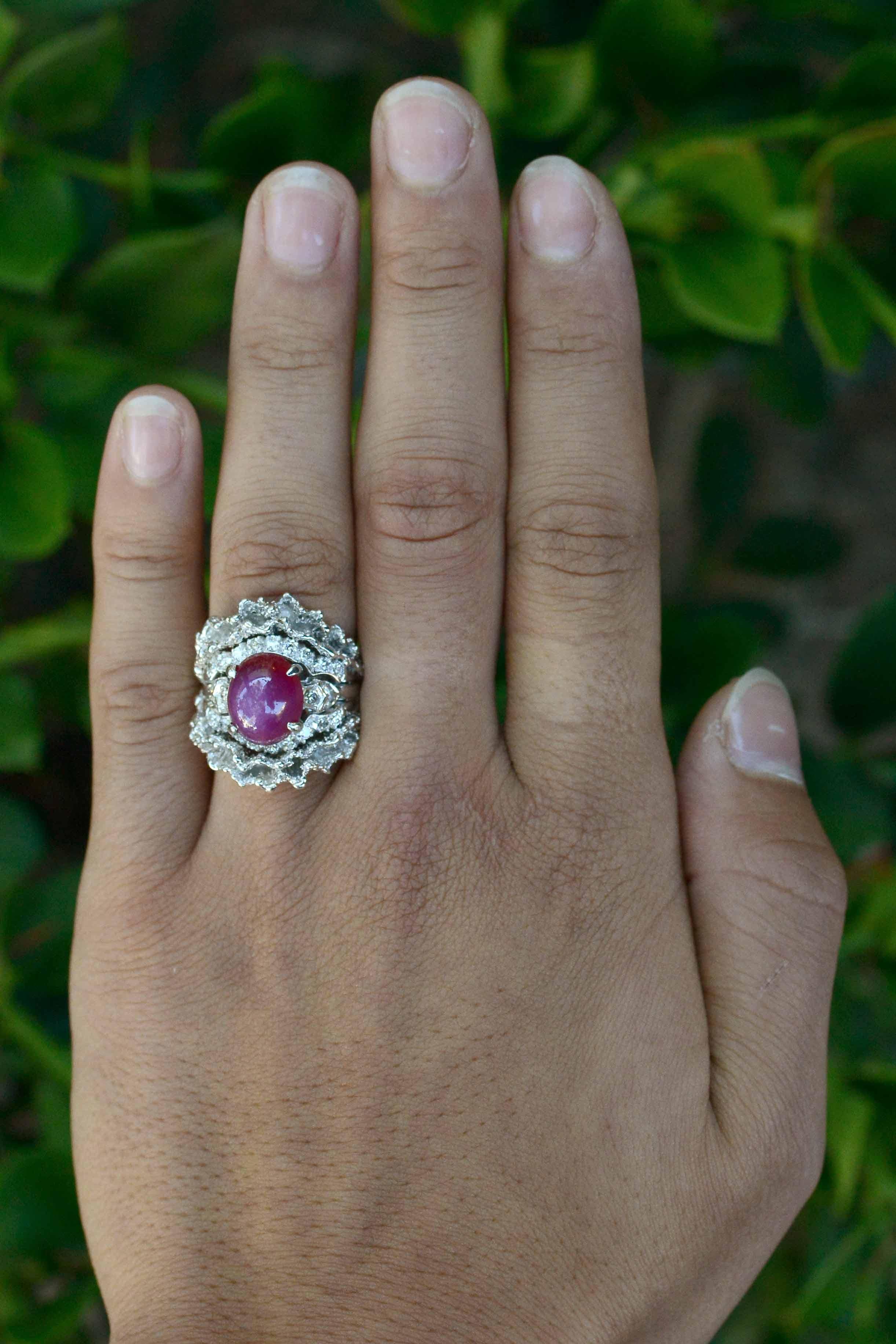A blazing 5.61 carat star ruby nestled in a dome setting adorned with 24 sparkling diamonds make this Brutalist era cocktail ring really pop. The wide band featuring a ruffled cowl of 14k white gold surrounds a most pleasing, vivid pinkish violet