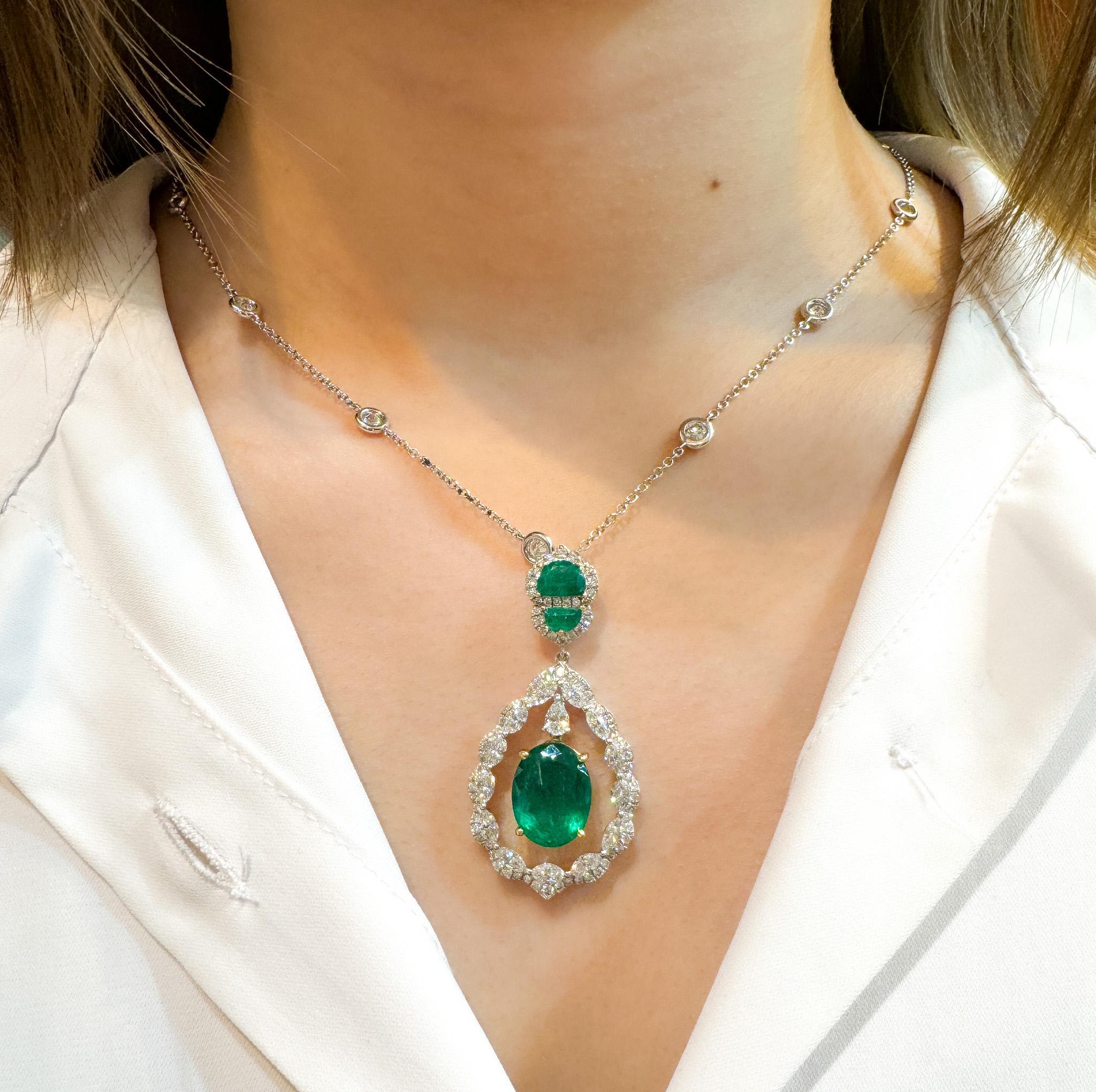 The natural oval-cut emerald center stone weighs 6.42 carats and features a floating design. It is paired with a halo of 13 marquise-cut white diamonds weighing 1.67 carats total, 1 drop-cut white diamond weighing 0.21 carats, and 80 round-cut white