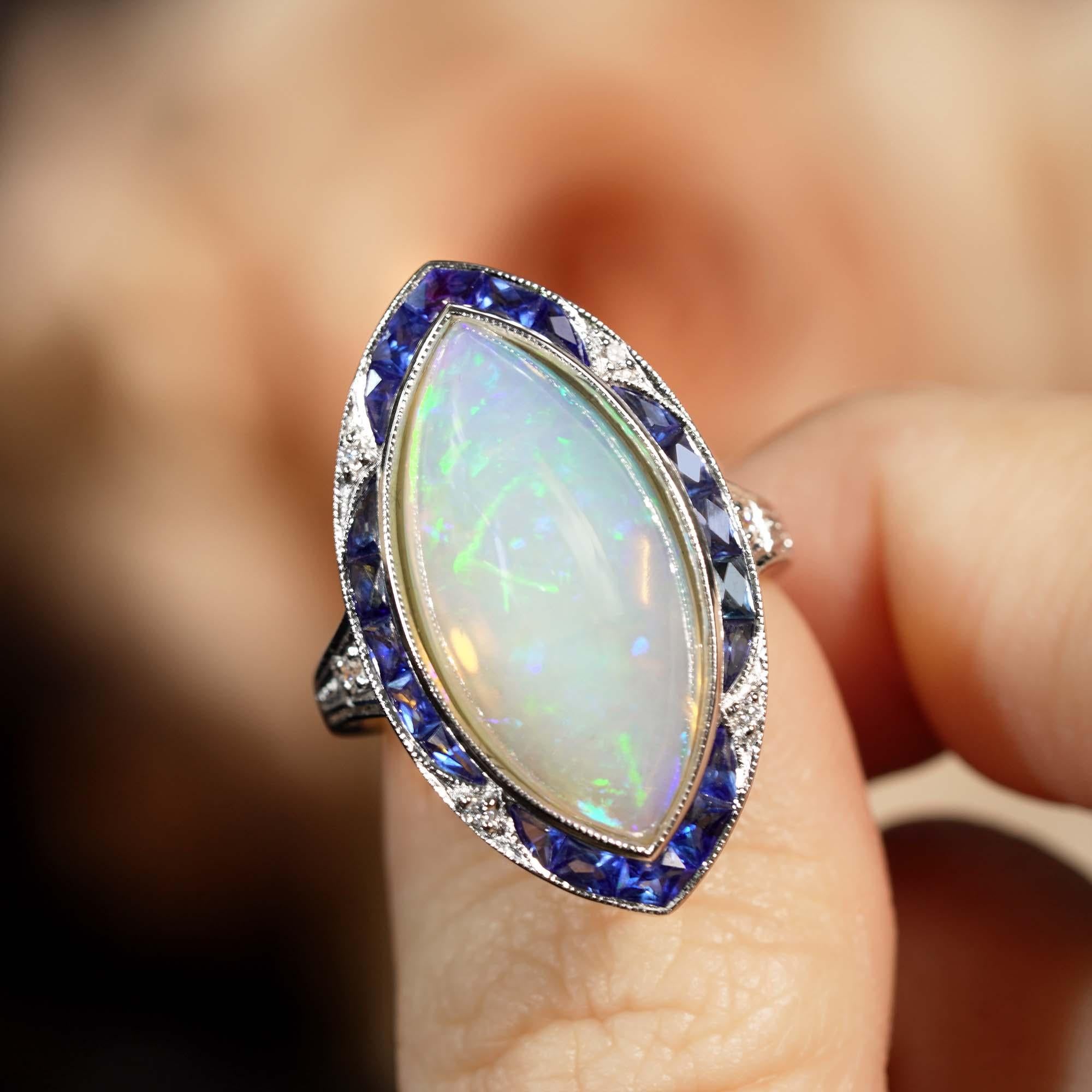 The soft milky white hue of marquise shaped cabochon opal that glows at the center of this alluring Art Deco inspired ring is punctuated throughout with iridescent flecks of green, orange, violet and blue and is set in a milgraning edged white gold