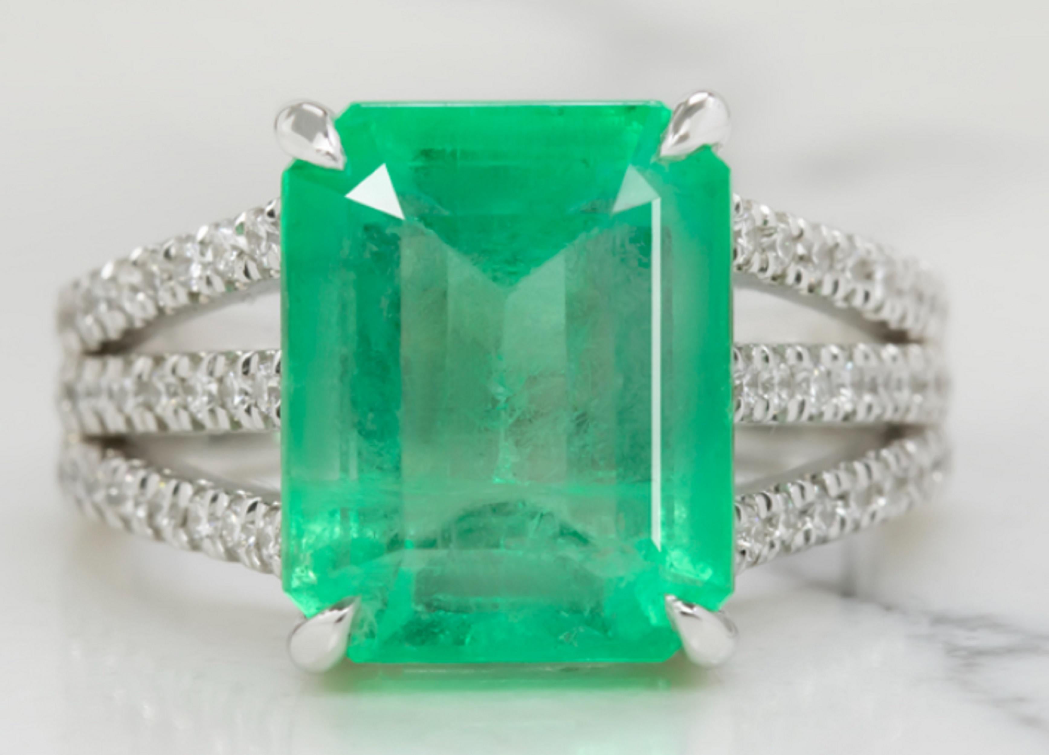The 6.80 carat emerald is a beautifully vibrant and highly saturated spring green hue, and its sophisticated emerald cut displays lovely sparkle. The claw prong setting offers a chic modern touch while the diamond encrusted triple split shank adds
