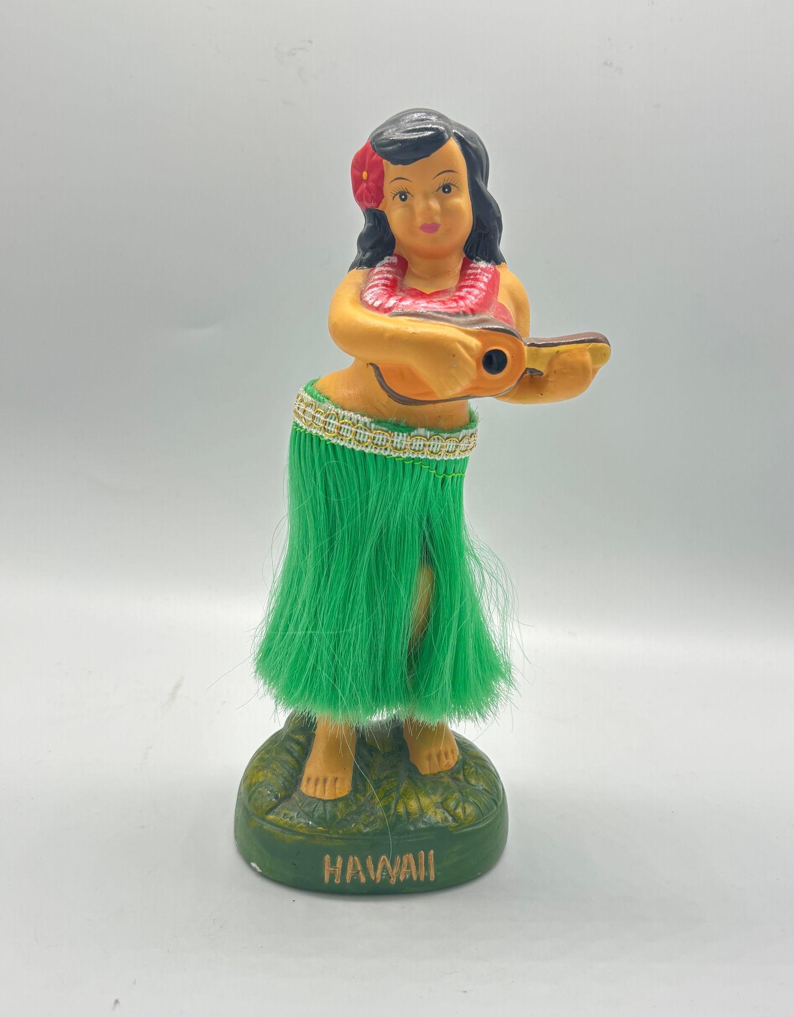 A Ceramic Hawaiian girl nodder figurine depiction of a young girl dressed in traditional Hawaiian attire, includes a colorful floral dress, a flower lei around her neck, and a flower in her hair. She is standing on a base with a spring-loaded