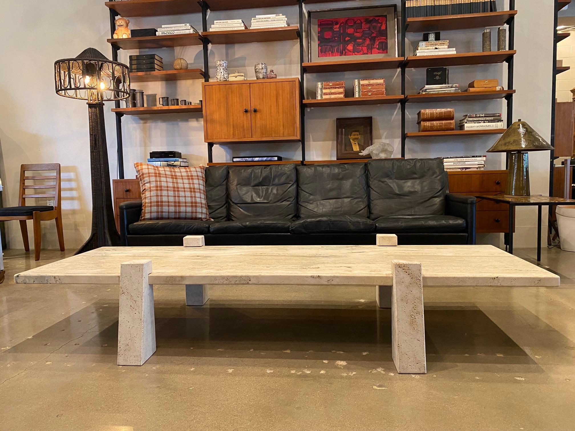 This is a monumentally impressive travertine cocktail table.  The thick monolithic honed slab top nestles into slotted angular post legs, whose position along the slab are adjustable.  All natural finished travertine posts subtly contrast with a