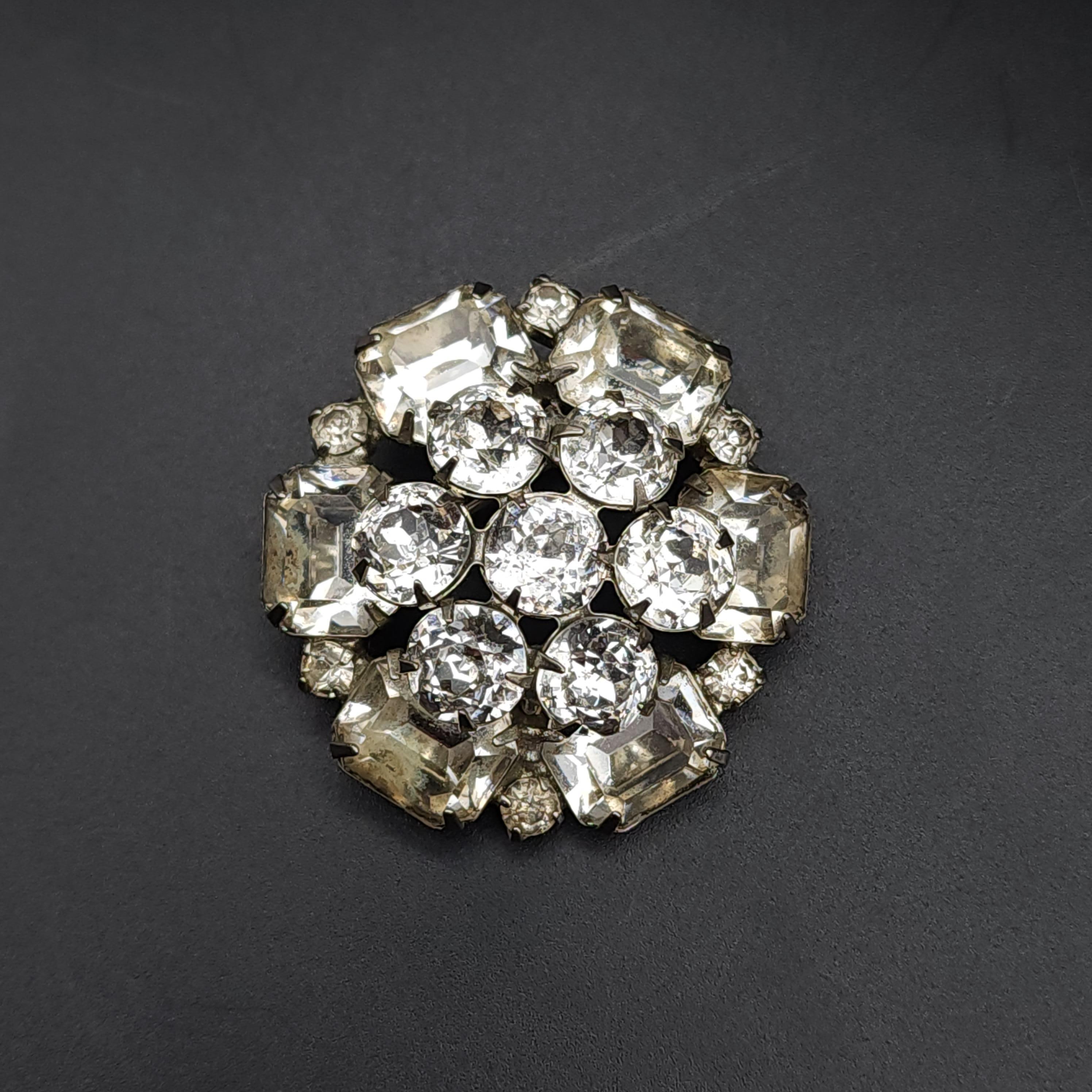 Größe: 4 cm / 1,5 in
Silber-Ton
Klare Kristalle

Mit dieser exquisiten, vom Art déco inspirierten Brosche aus den 1970er Jahren werden Sie in die Vergangenheit zurückversetzt. Diese runde Brosche präsentiert eine Reihe von klaren Kristallen im