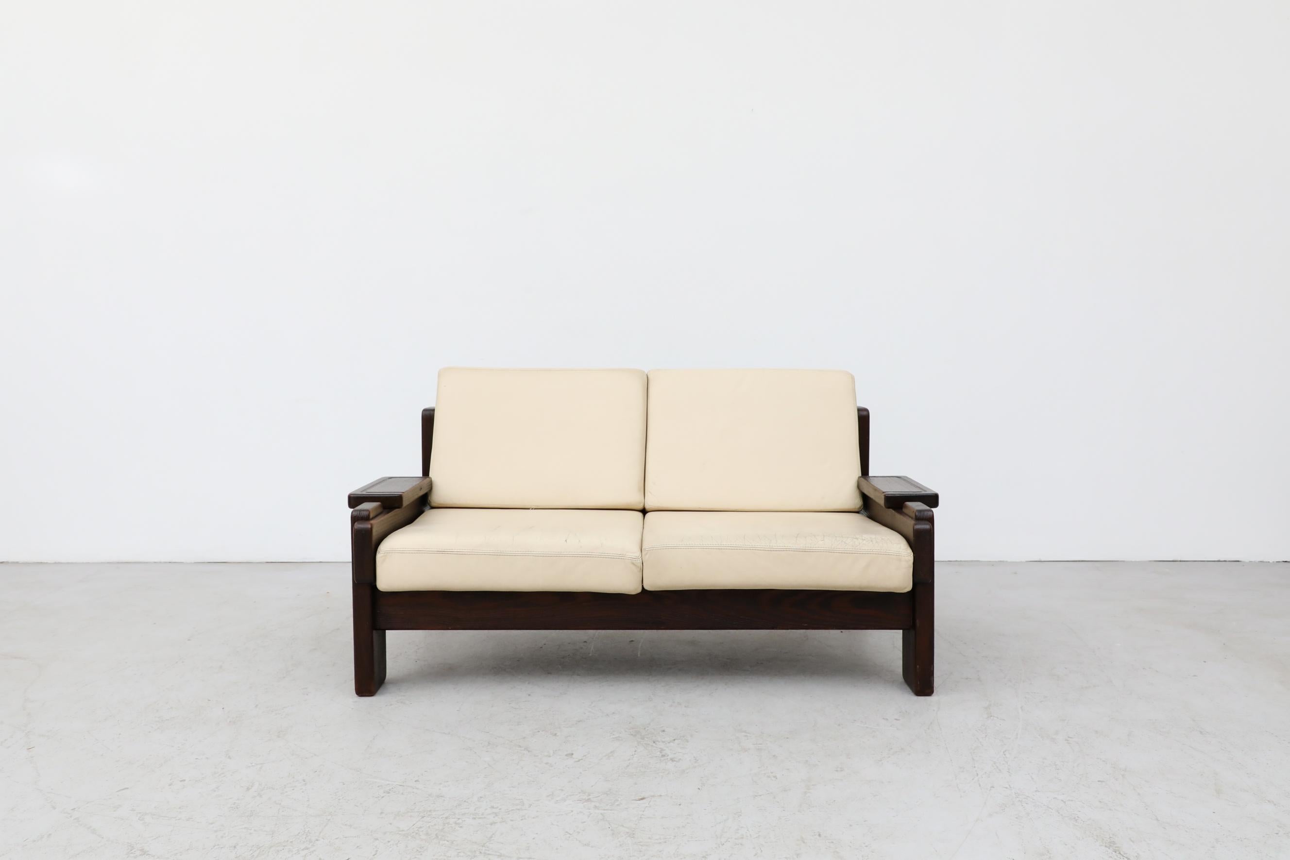 70's Brutalist leather loveseat with cream cushions and a dark heavy wood frame. In original condition with visible wear to the leather, consistent with its age and use. A matching 3 seater sofa is available and listed separately (LU922428505952).