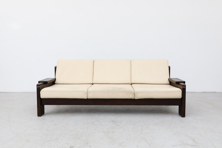 70's Brutalist leather sofa with cream cushions and a dark heavy wood frame. In original condition with visible wear to the leather, consistent with its age and use. A matching loveseat is available and listed separately (LU922428493242).