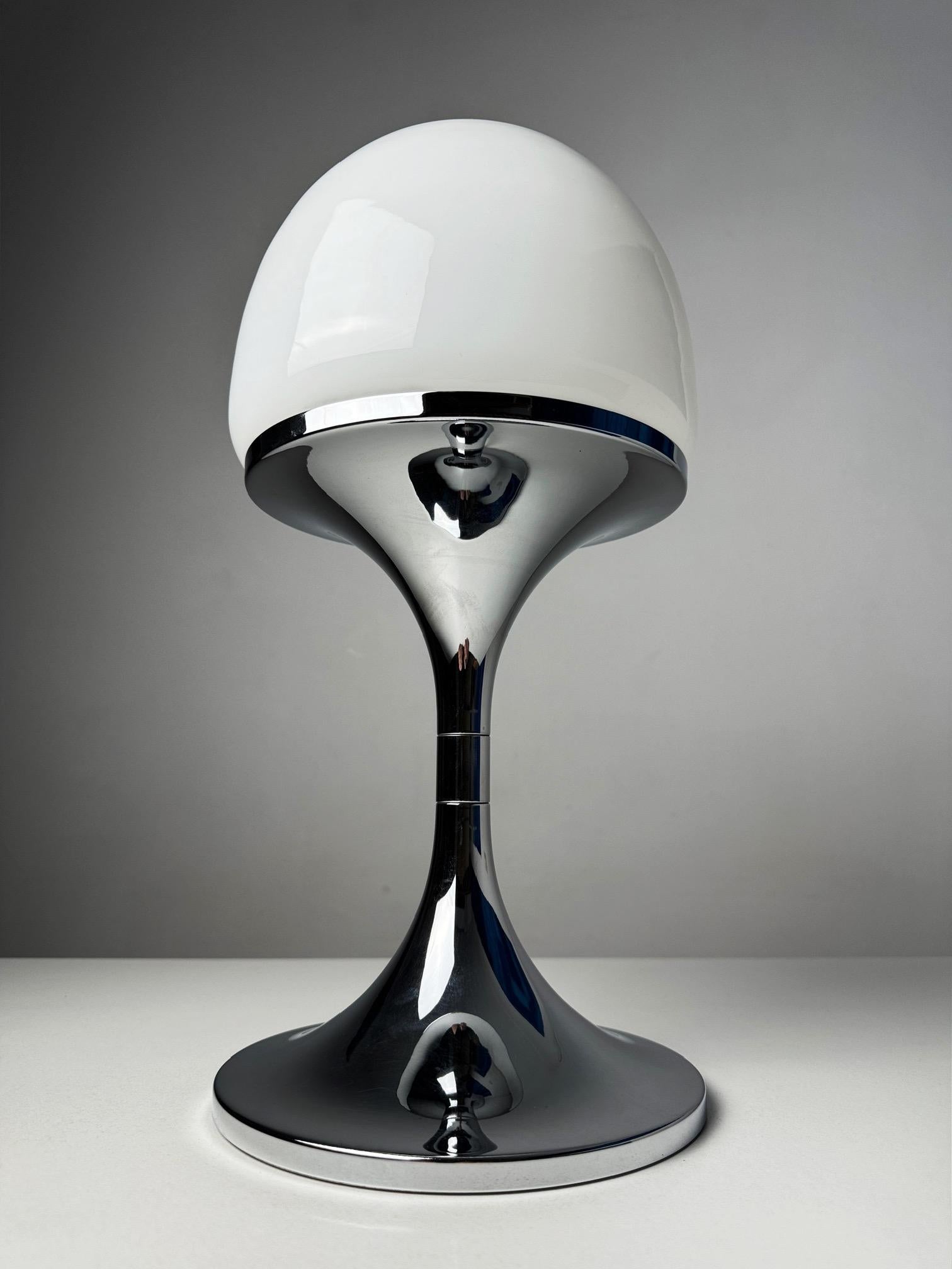This elegant table lamp from the 1970s embodies the distinctive aesthetics of the Space Age era with its sleek chrome base and soft opaline glass shade. The design features clean lines and a minimalist silhouette, making it a perfect statement piece