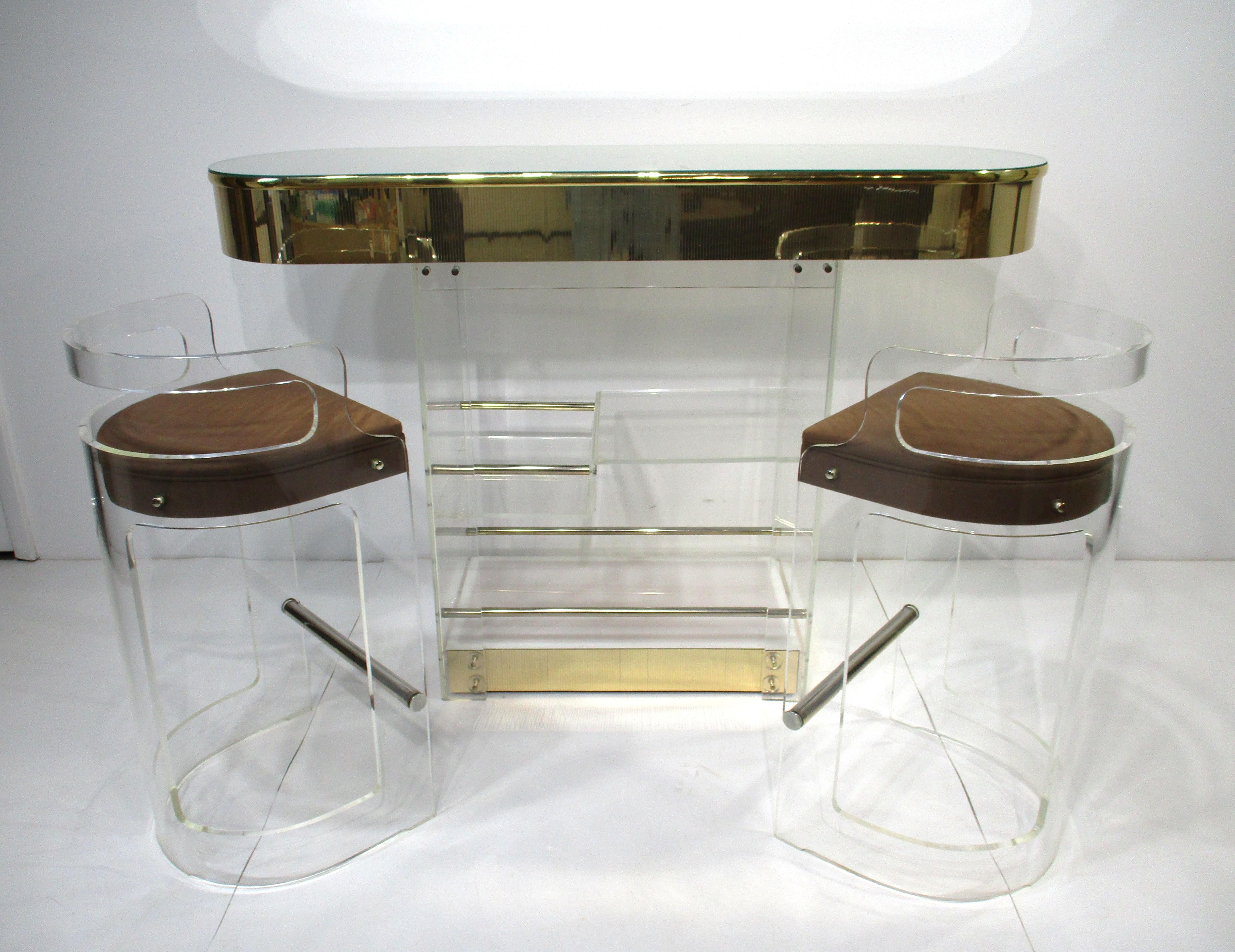 A luxurious and stunning three-piece bar set in Lucite with mirrored top, gold tone trim and matching upholstered bar stools in the manner of Charles Hollis Jones. The back of the bar has bottle holders built into the top and the lower area has