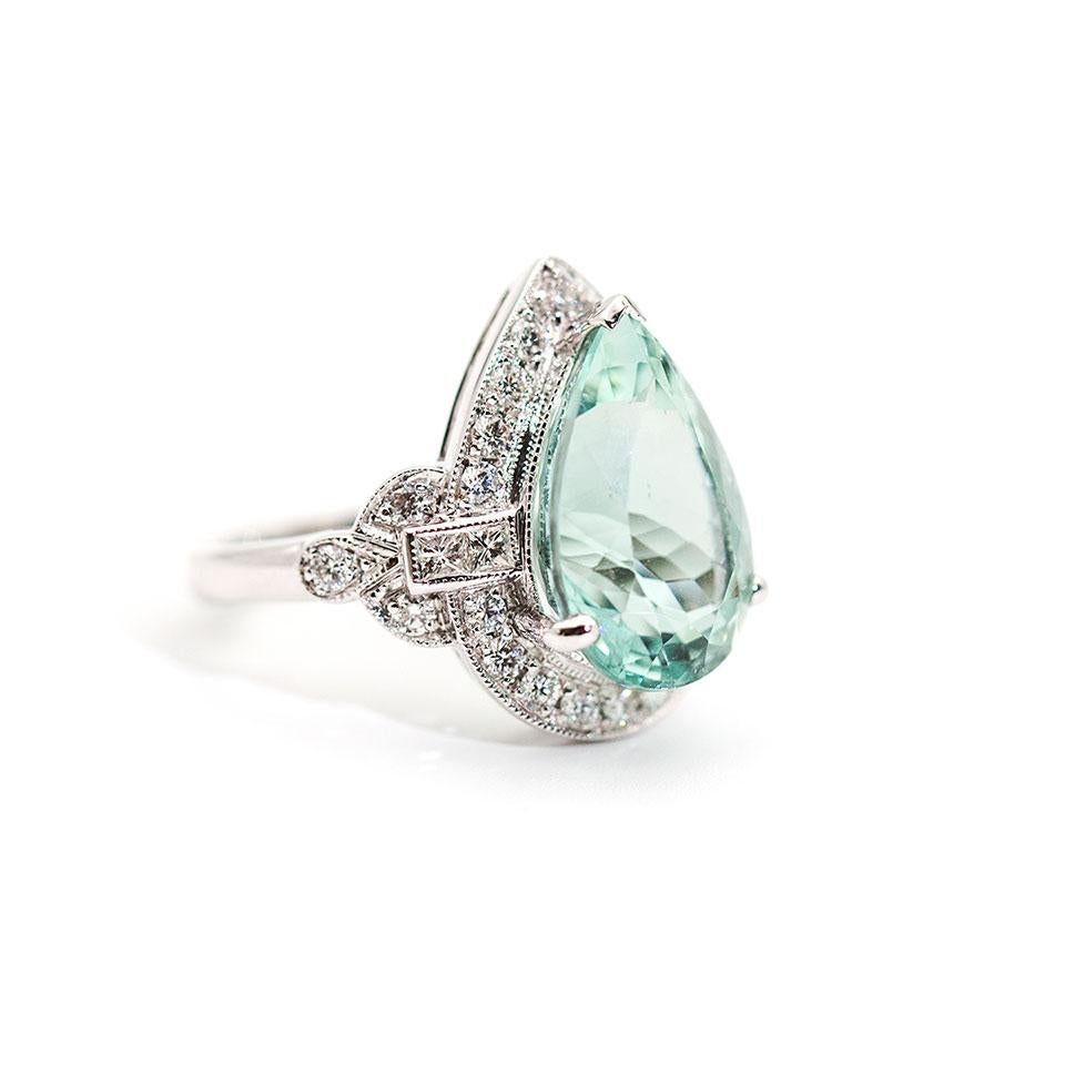 This striking art deco inspired designed ring is forged in 18 carat white gold and boats a captivating 7.47 carat flawless bright bluish green pear cut natural Aquamarine surrounded by 0.87 carats of alluring round brilliant cut diamonds and