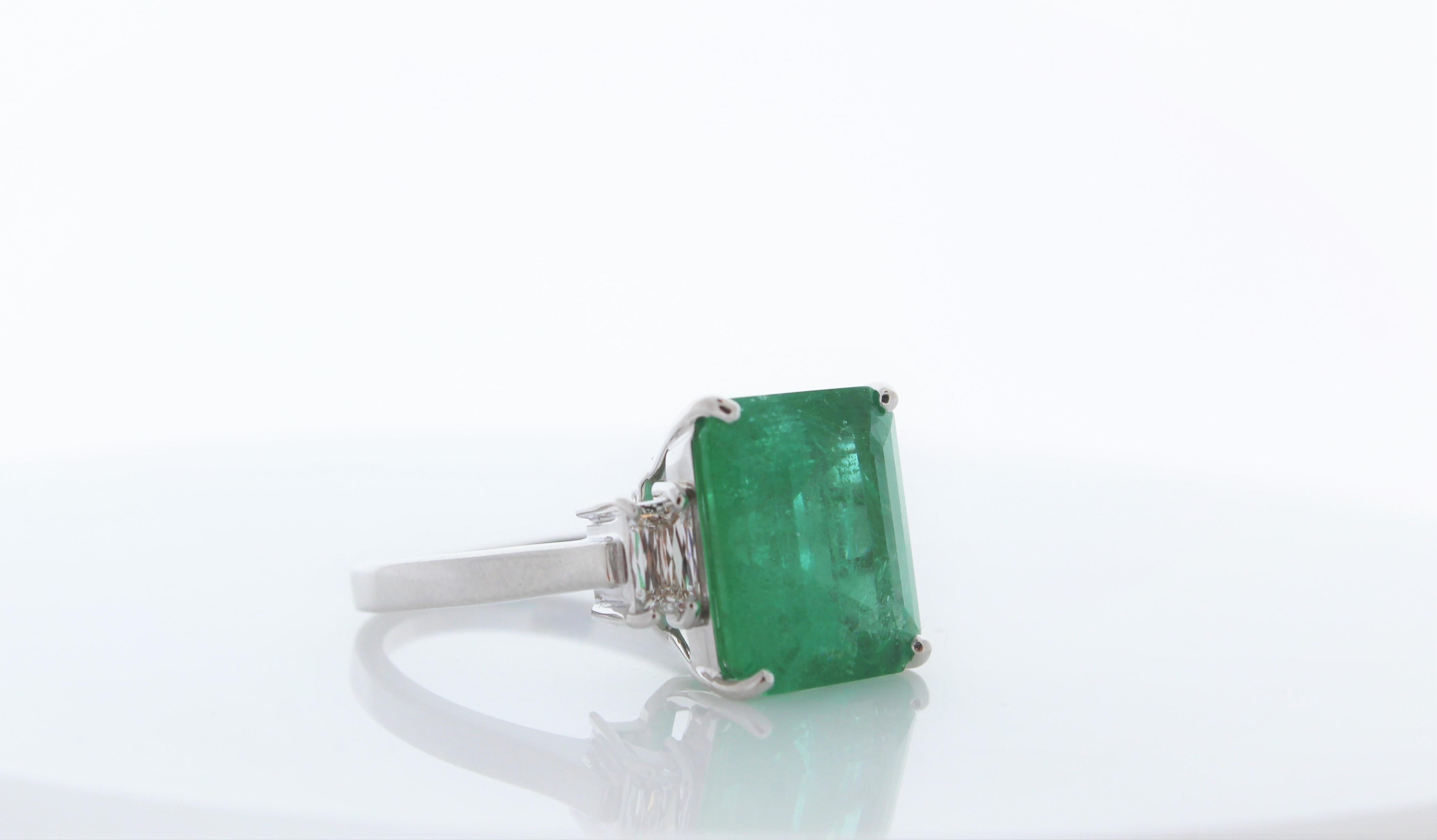 This is a 7.70 cushion cut intense green emerald. The gem source is Colombia; its color is vibrant grass green. Its transparency and luster are excellent. There are two diamonds totaling up to .84 carats. Created in brightly polished 18 K white