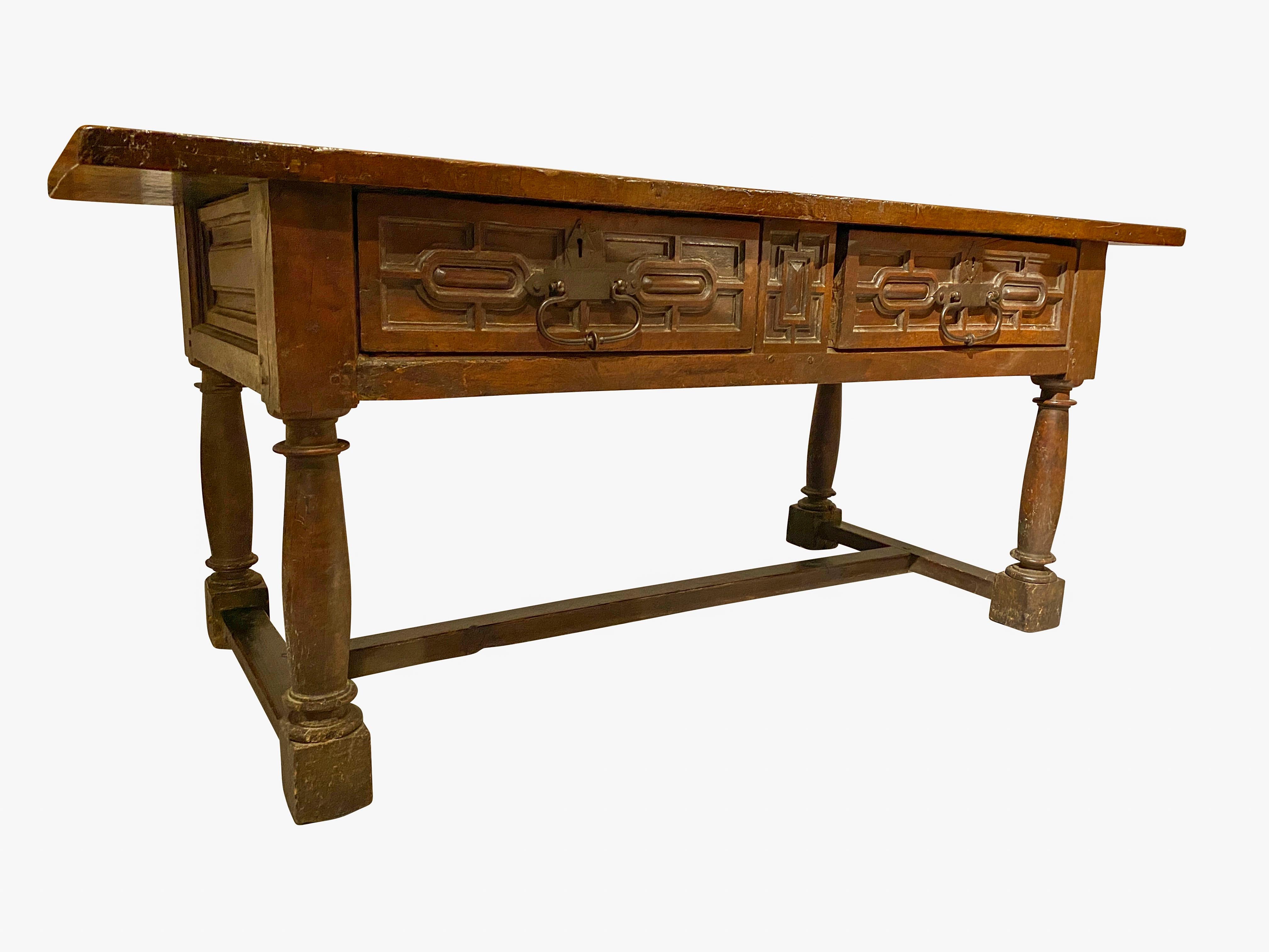 With a thick single board top over two carved panelled drawers flanking a geometric form panel, the drawers with nicely wrought iron handles and escutcheons, panelled sides, raised on turned legs joined by an H form stretcher.