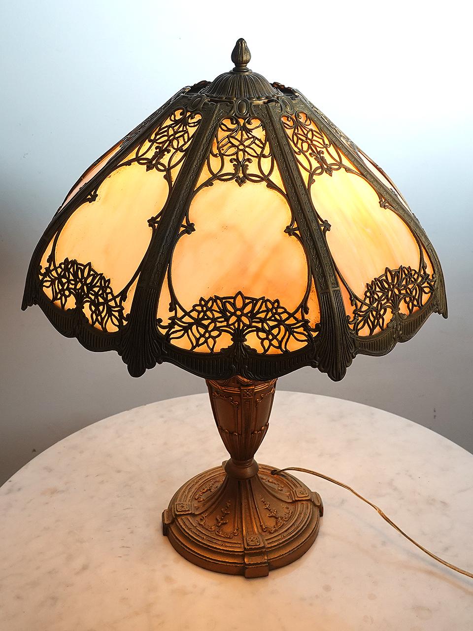 This is a nicely designed Arts and Crafts table lamp. Its impressive at 2 foot tall with a 19 inch diameter. It has a curved shade with 8 cast floral filigree panels. Each slump glass (curved) panel features a cream -  amber variegated glass and is