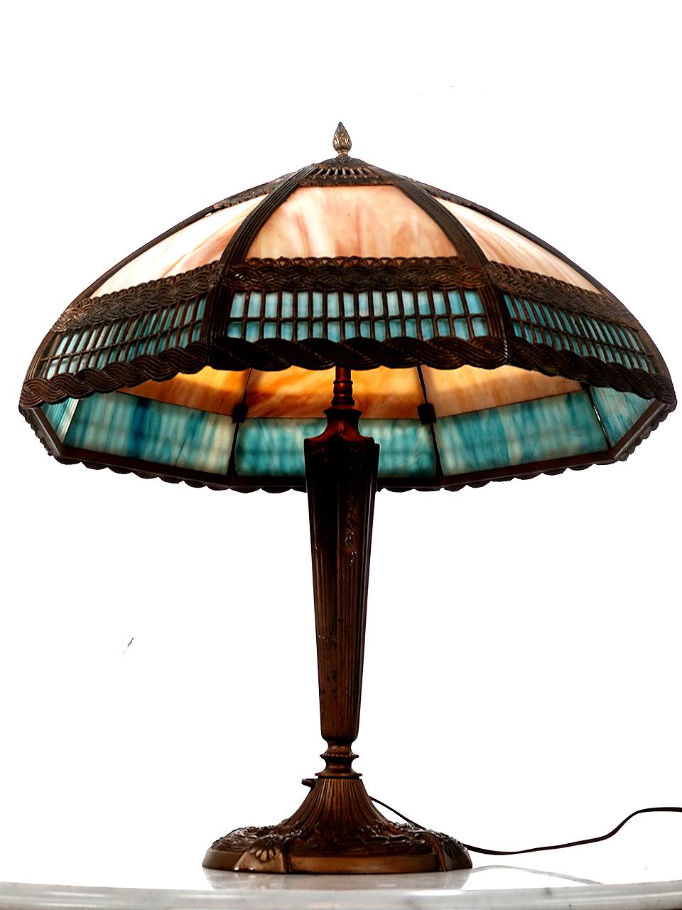 This is a nicely designed Arts and Crafts table lamp. Its impressive at 2 foot tall with a 21 inch diameter. It has a curved shade with 8 cast filigree panels. The design has an interesting tied stick and rope theme. Each slump glass (curved) panel