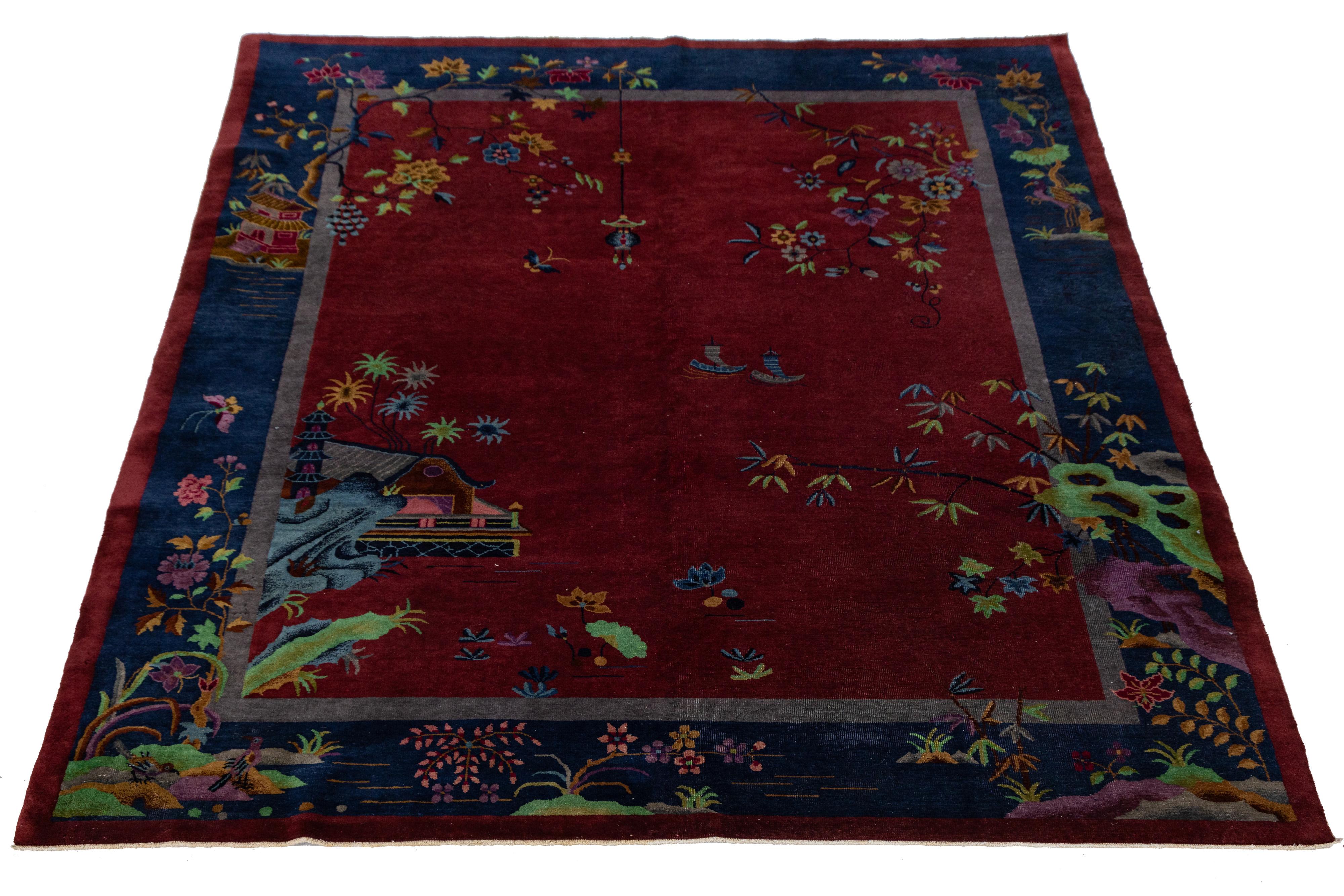 Ce tapis chinois antique en laine des années 1920 présente un champ rouge vif encadré par des bordures indigo profondes, mettant en scène un paysage paisible avec des pagodes, des bateaux, des cascades et des motifs floraux luxuriants. Tissé à la