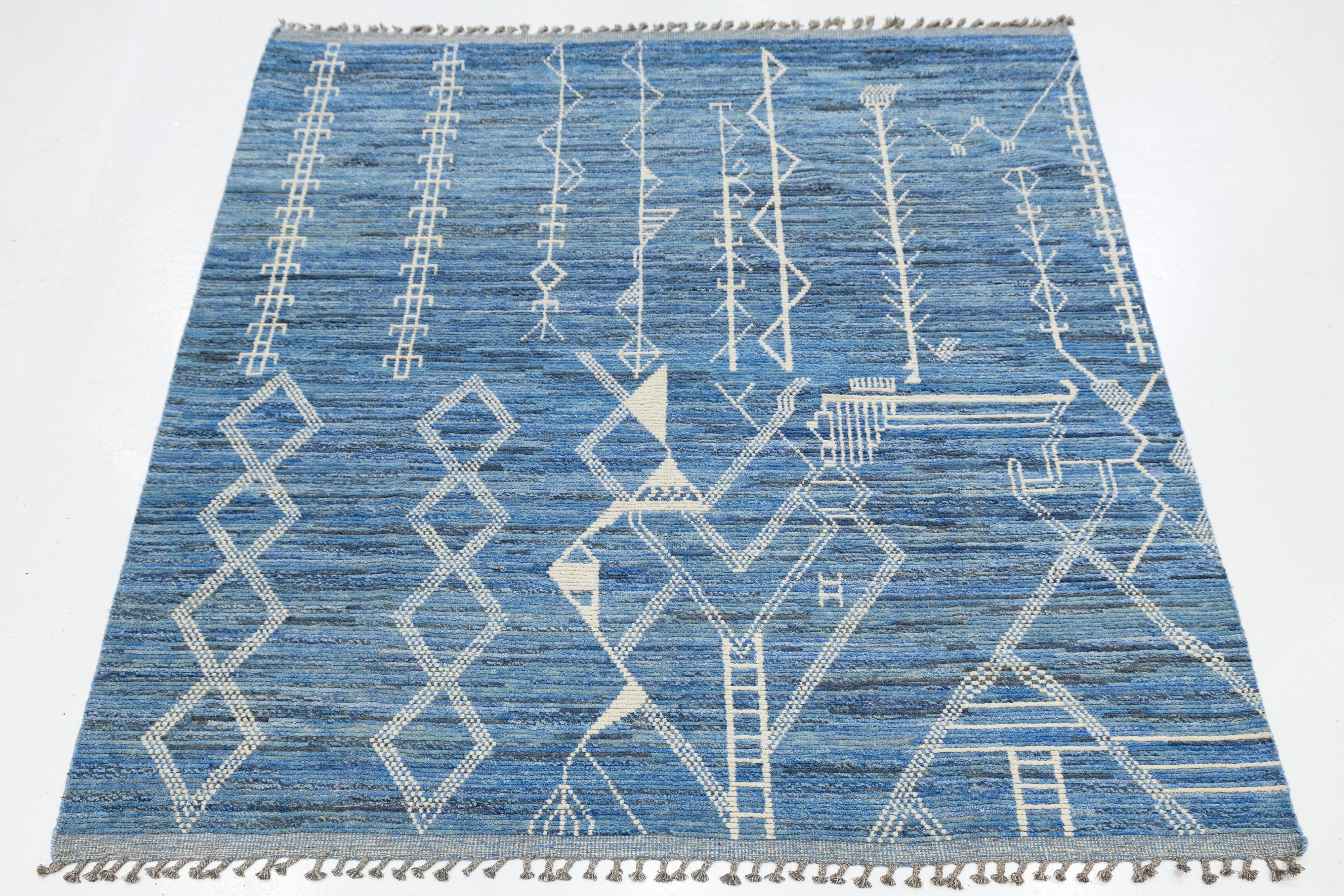 This modern Moroccan-style wool rug features intricate white geometric and symbolic motifs over a rich blue field, blending tradition with contemporary artistry. Handwoven with fringe edges, its bold design and layered texture make it a striking