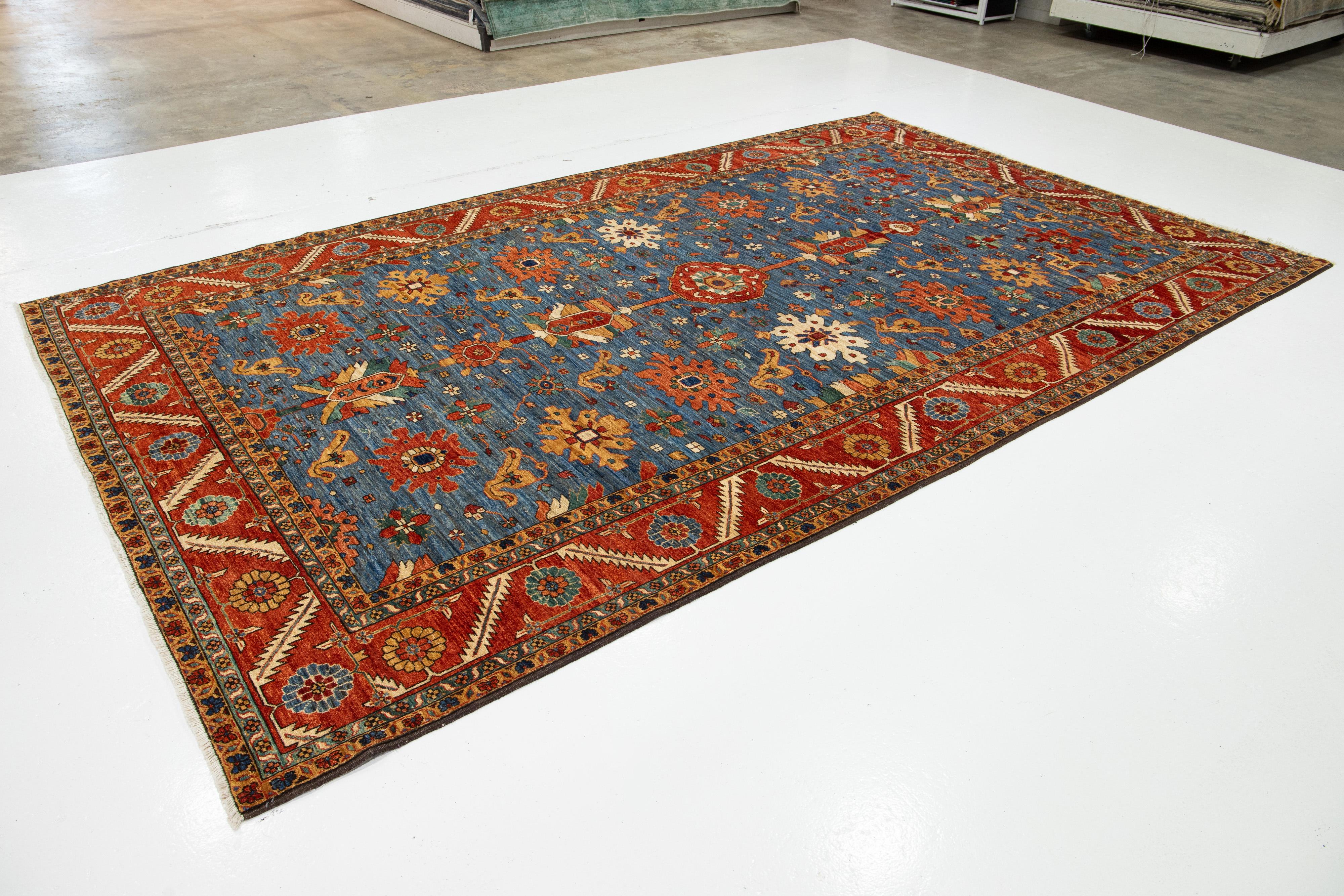 This refined wool rug features a timeless blue backdrop paired with a contemporary Serapi-inspired design. The piece showcases a red frame and a stunning all-over floral pattern adorned with exquisite multicolor accents. It brings sophistication and