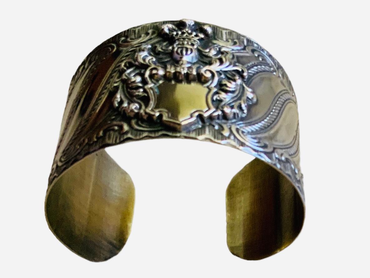 This is an 800 silver repousse cuff bracelet . It is decorated in the center with a shield. This shield depicts a bust of a knight armor. Over its helmet, there is crown with the Prince of Wales feathers. Acanthus leaves embellish also it at both