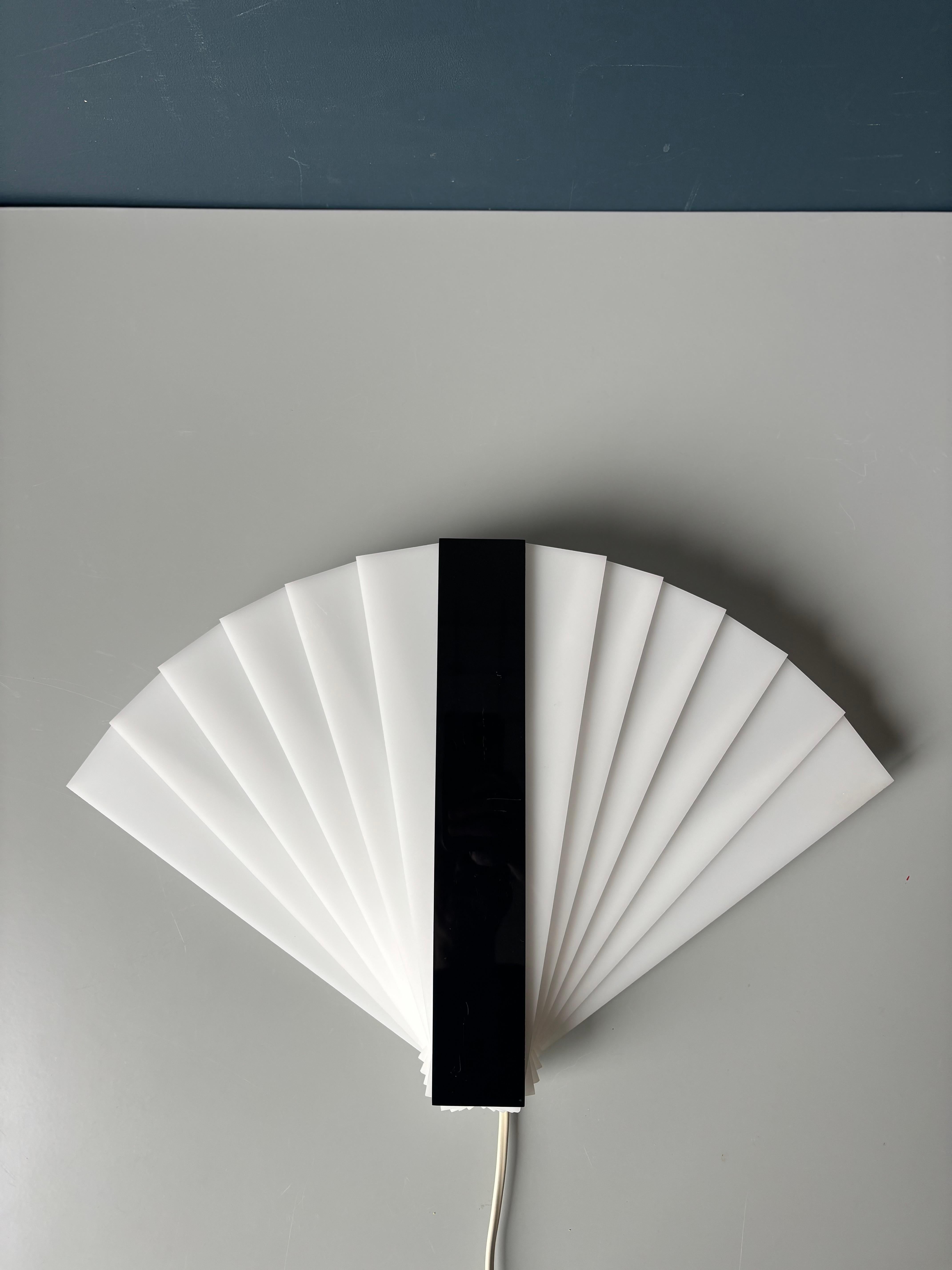 Lampada da parete ispirata all'Art Déco, composta da 12 sottili bastoncini acrilici bianchi a forma oblunga posizionati in successione dietro un unico bastoncino acrilico nero per creare una grande forma a ventaglio. Le dimensioni di questa lampada