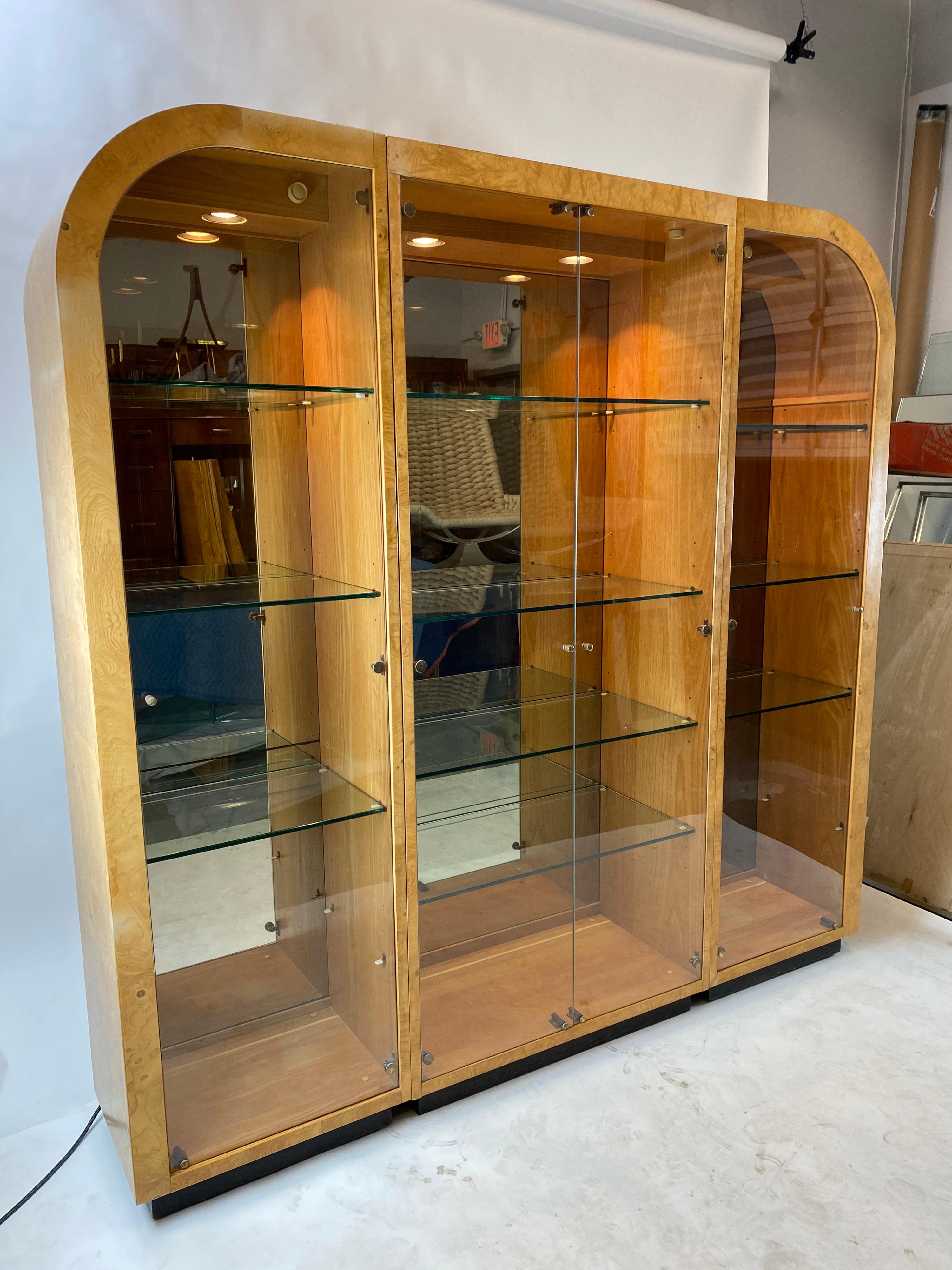 A timeless, 80s modern, three-piece Henredon display cabinet featuring a beautiful maple burl exterior, ebonized base, mirrored interior back panels, and lighted interior featuring thick glass shelves. Each of the three units are lighted with