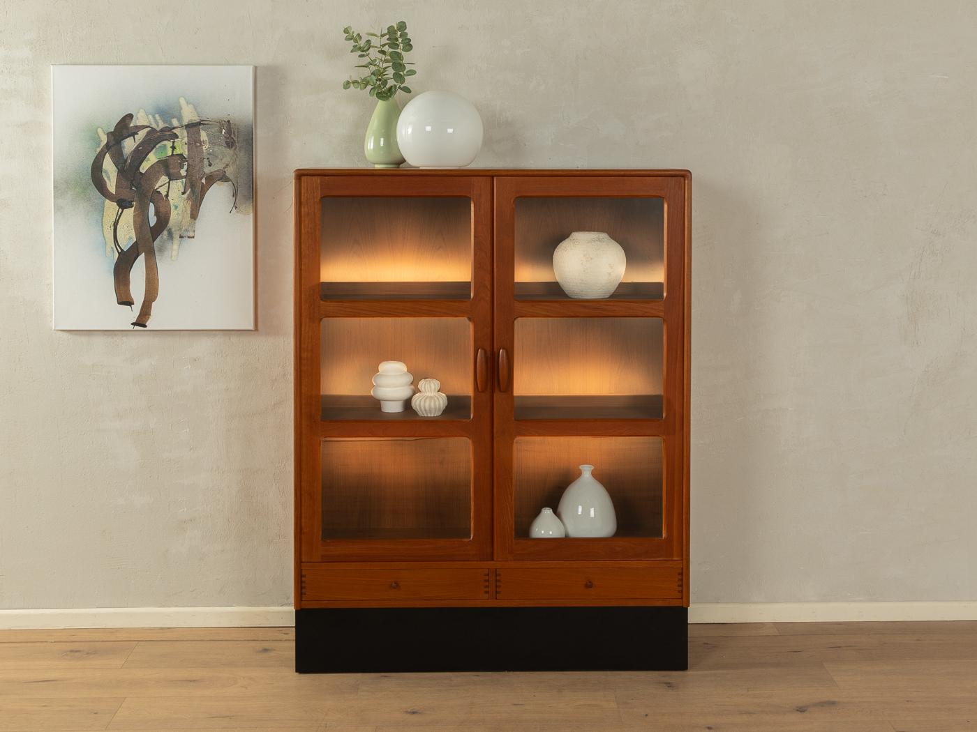Classic showcase from the 1980s. High-quality corpus in teak veneer with two glass doors, two shelves, two drawers and a black contrasting base. The showcase can be illuminated.

Quality Features:
very good workmanship
high-quality materials
corpus