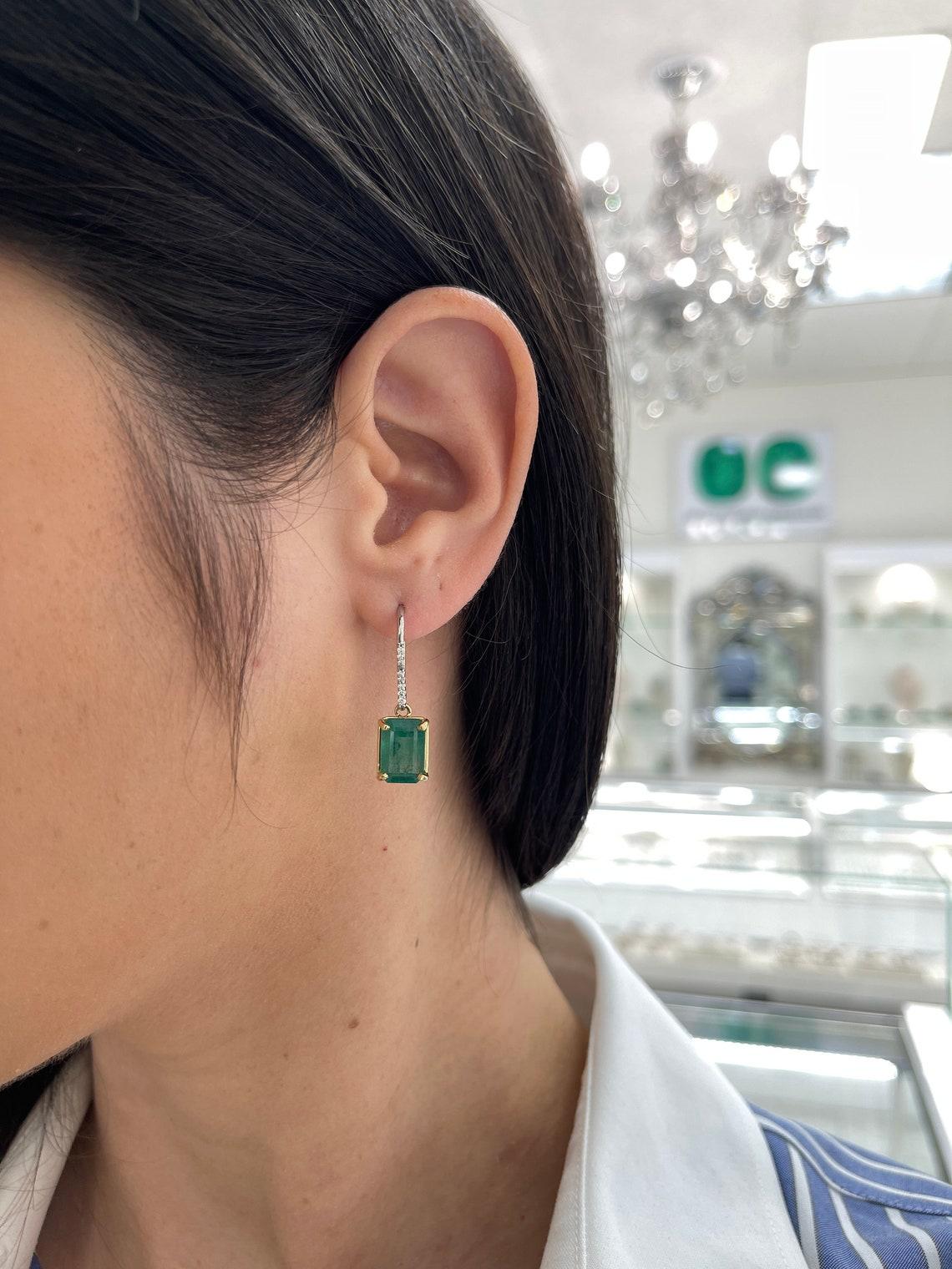 Élevez votre élégance avec nos boucles d'oreilles pendantes en émeraude et diamant, mettant en vedette des émeraudes taillées en émeraude d'une luxuriante couleur vert moyen foncé et d'une clarté semi-transparente. Ces pierres précieuses captivantes