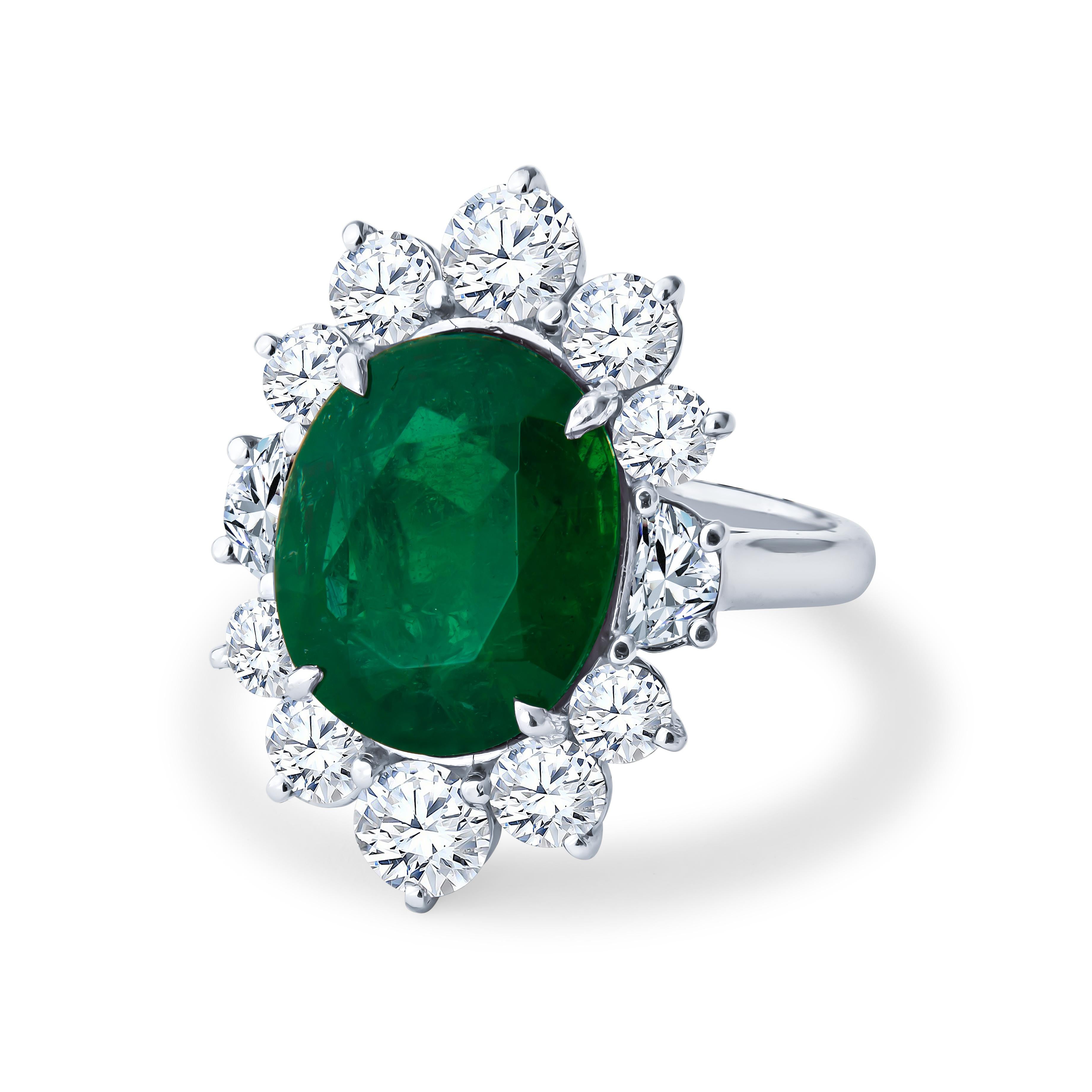This exquisite colored stone piece features a 8.44ct oval cut emerald, surrounded by a halo of 3.35ct total weight in 10 round cut diamonds and 2 half-moon step cut diamonds, creating a floral design. The emerald center stone is a rich bright green