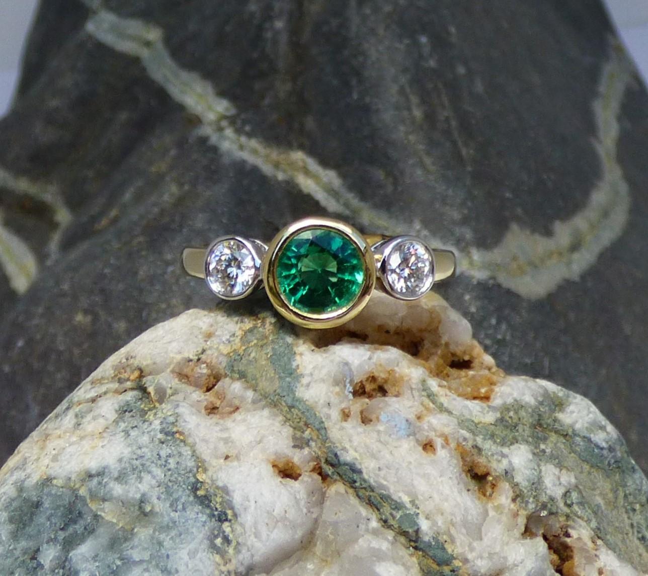 An exceptionally bright and colourful Emerald is set between two Diamonds. The Emerald is 7mm round and .85ct. in weight. The two Diamonds have a total Diamond weight of .43ct.  The stones are bezel set in a handmade 18K yellow ring with the