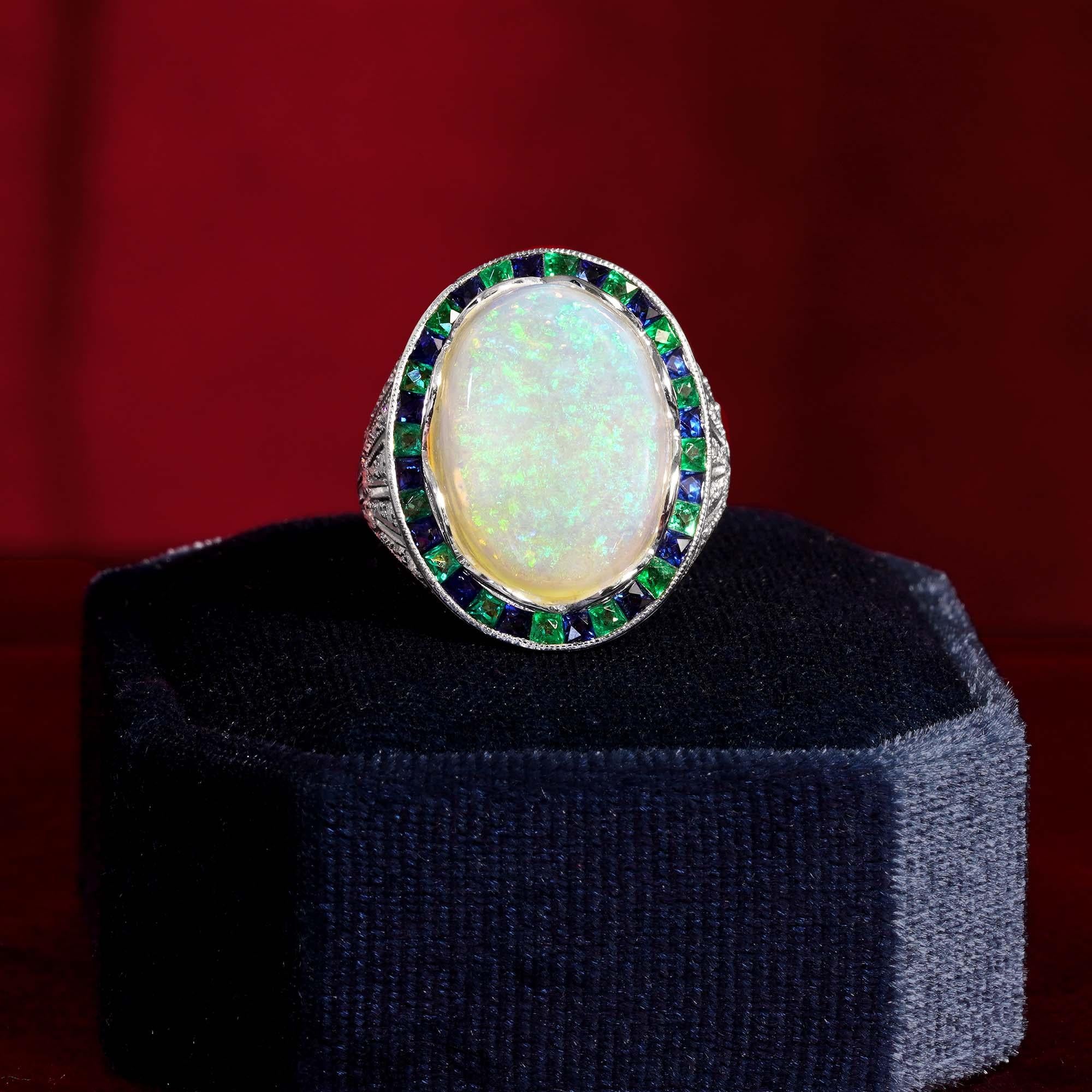 Radiating vintage charm, this Art Deco style cocktail ring features a luminous opal at the center.Encircled by vivid French-cut emeralds, the design glows with color and elegance.Diamond-accented shoulders add sparkle and sophistication to every