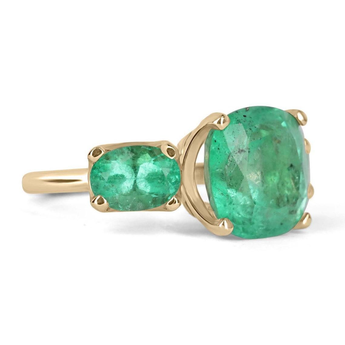 A Colombian emerald three stone ring. This bespoke ring design is crafted in gleaming 14K yellow gold, this ring features a 6.55-carat cushion cut in its center. Oval emeralds are flanked on the side creating an array of green. Simply gorgeous and