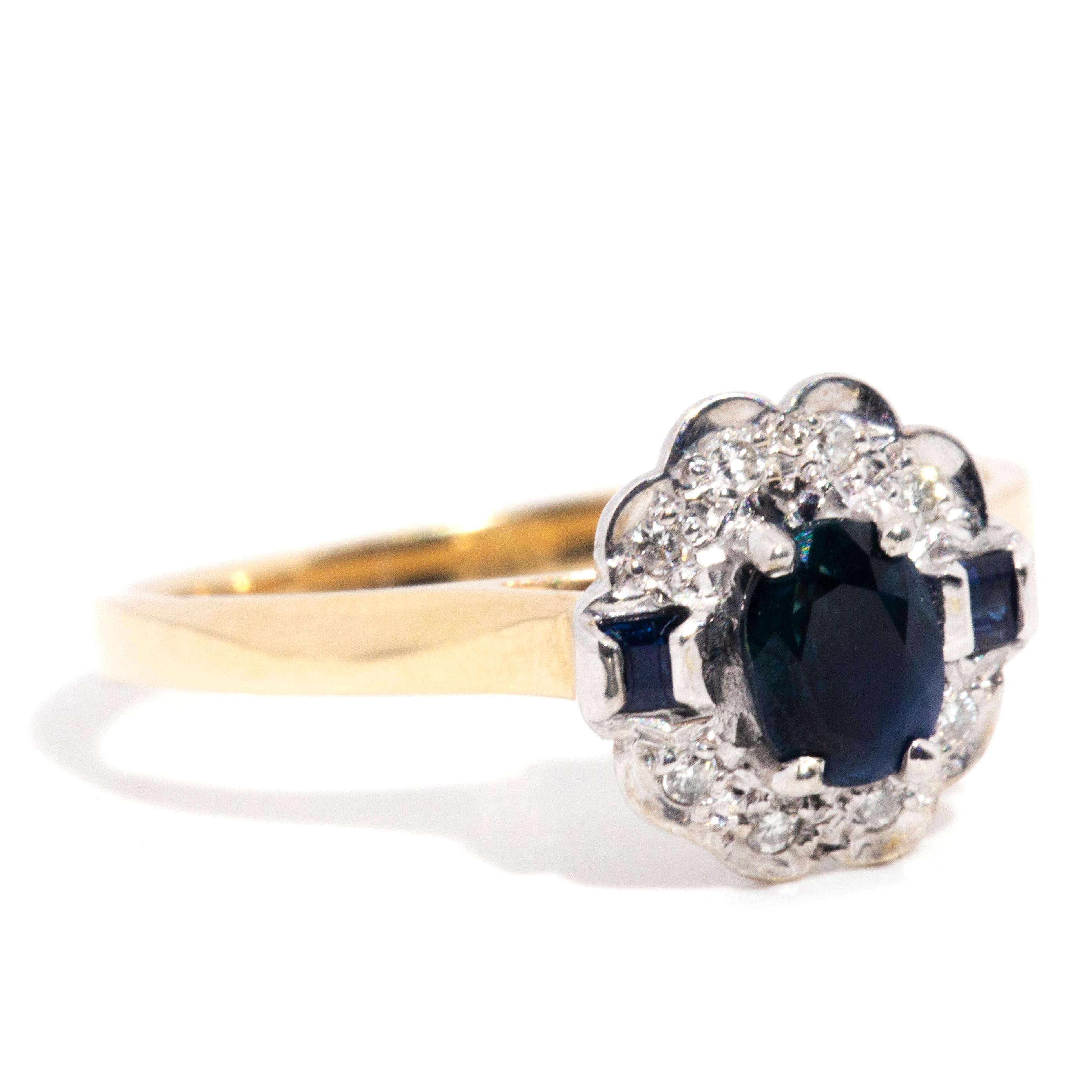 Forged in 9 carat yellow gold, this elegant vintage ring sees a charming parallel band rising to a basket gallery embellished with a gorgeous dark blue oval faceted sapphire at the centre. The surrounding halo cluster is made of round shimmering