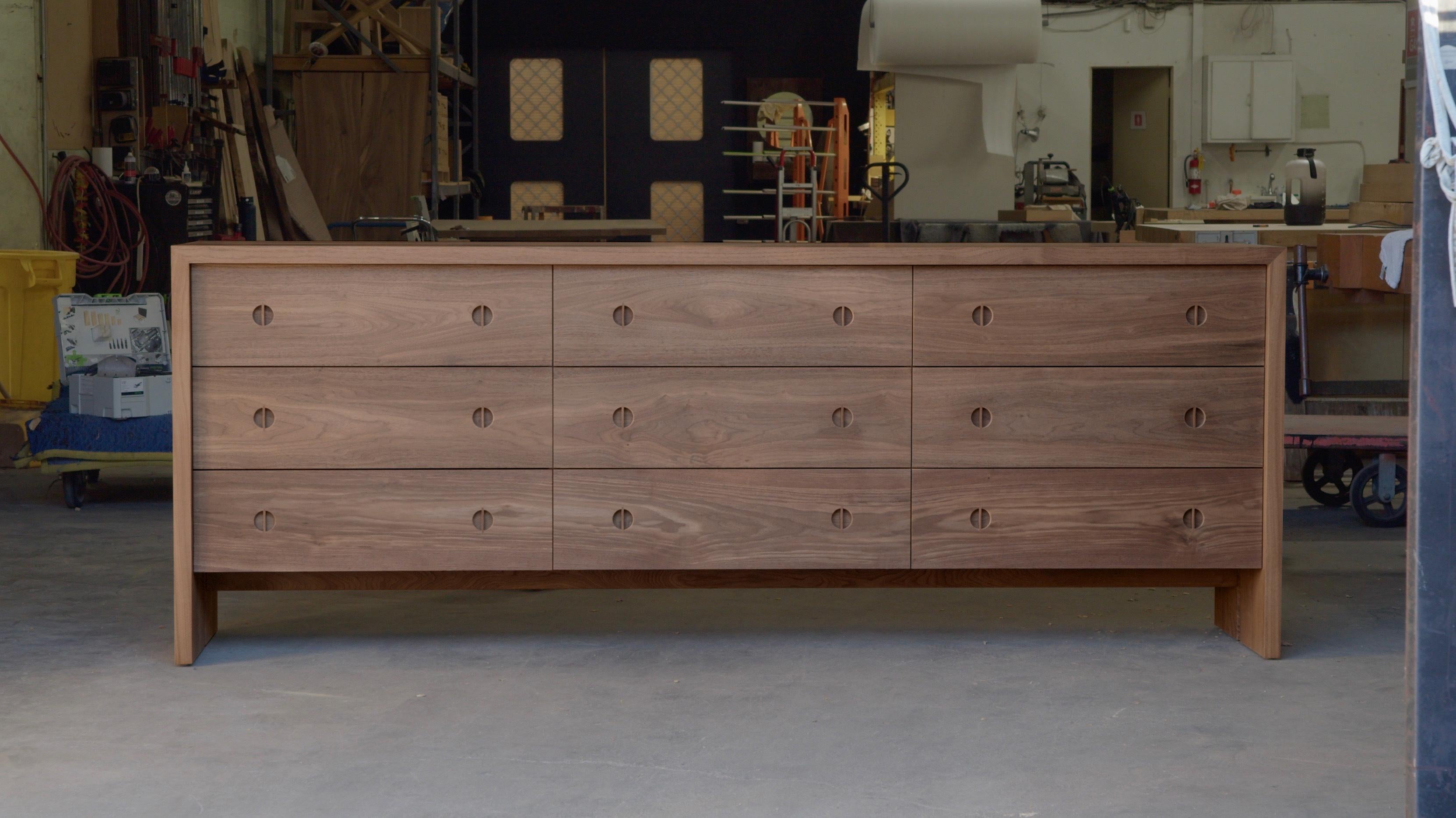 Questo comò moderno in legno è realizzato a mano negli Stati Uniti con una struttura interamente in legno duro. Caratterizzato da un corpo in noce massiccio, da un intarsio minimalista in Oak e dall'esclusivo dettaglio della gamba spaccata, questo