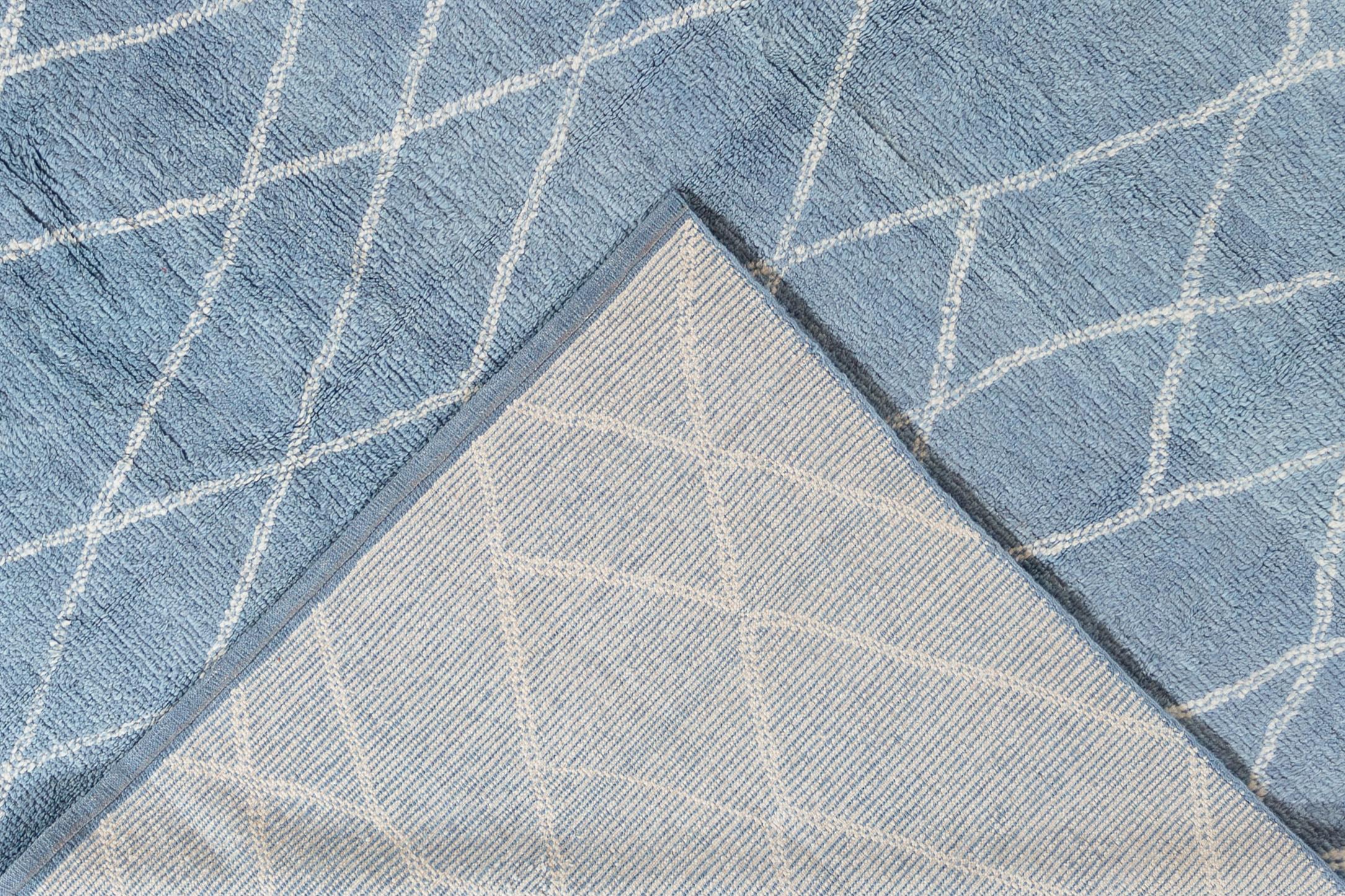 Experimenta la elegancia de esta alfombra contemporánea de estilo marroquí, con un suntuoso pelo de felpa anudado a mano con lana de primera calidad. La alfombra muestra un sereno campo azul claro adornado con un llamativo diseño tribal blanco, que