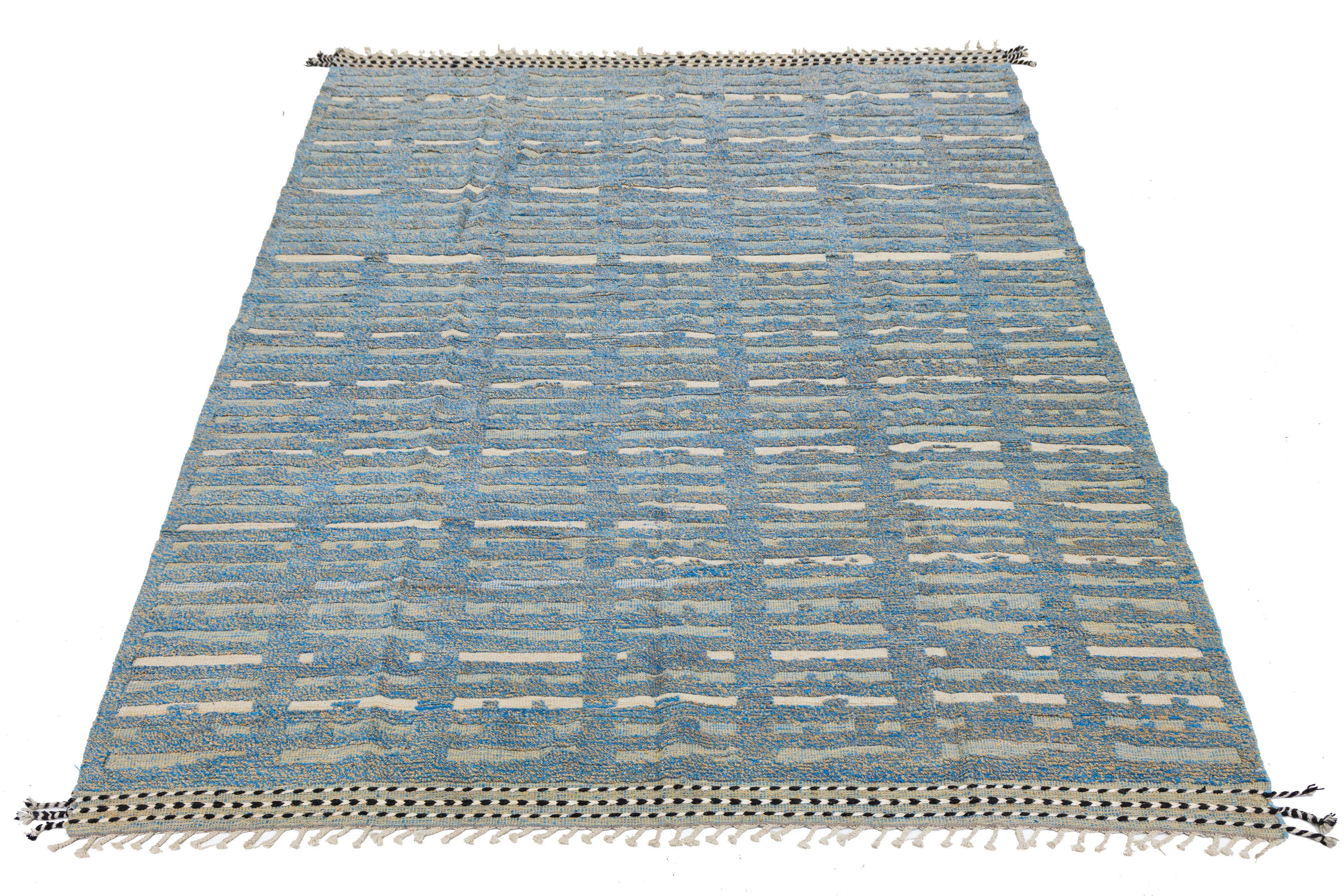Esta preciosa alfombra de lana presenta un diseño marroquí contemporáneo, resaltado por un elegante fondo tostado y beige. La moderna composición incorpora una llamativa mezcla de azules, creando un convincente impacto visual que sin duda captará la