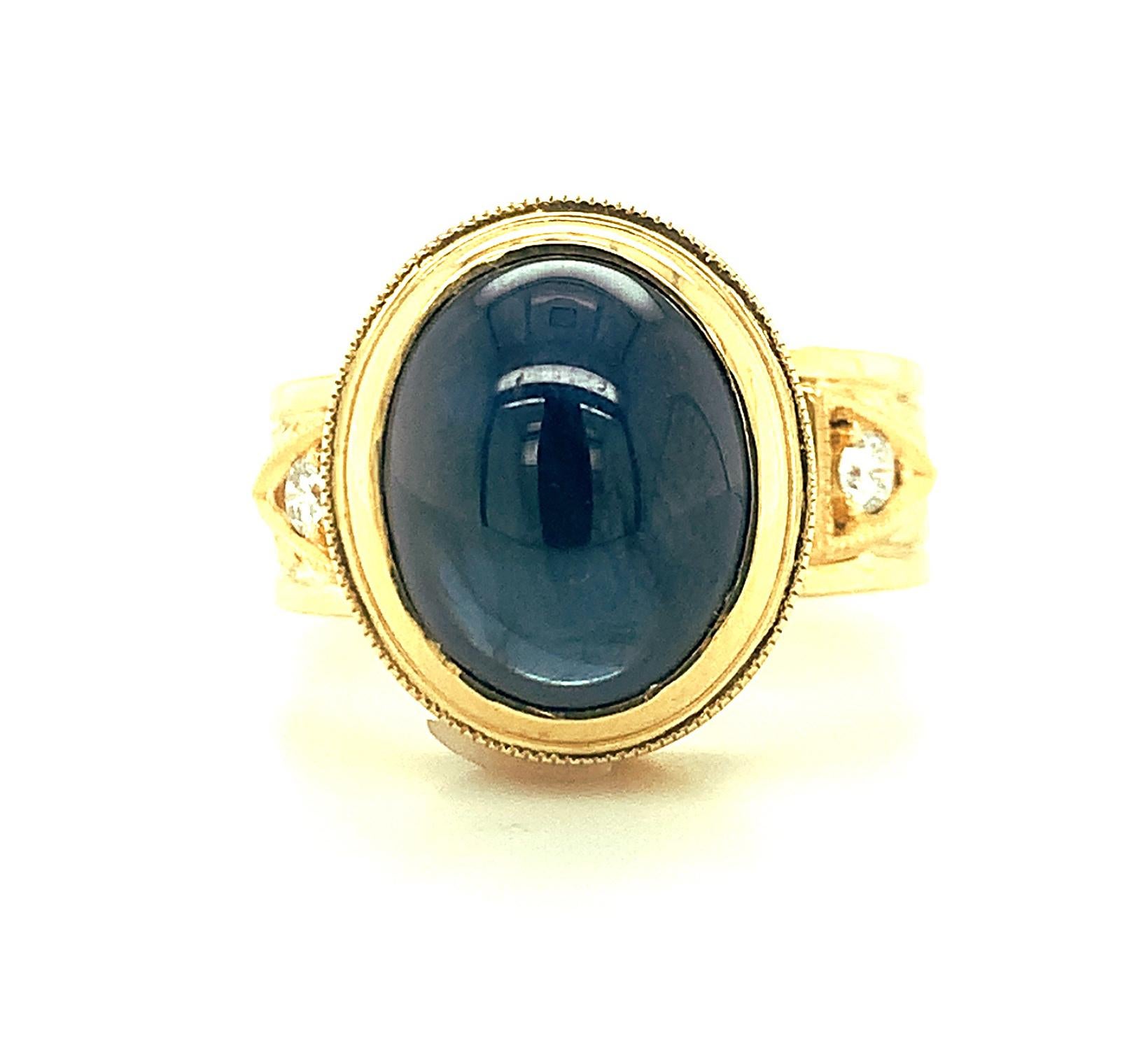 This handsome ring features a large, 9.06 navy blue sapphire cabochon, beautifully bezel set in an intricately engraved 18k yellow gold ring. The gorgeous hand engraved band displays stunning artistry and is set with two round brilliant white